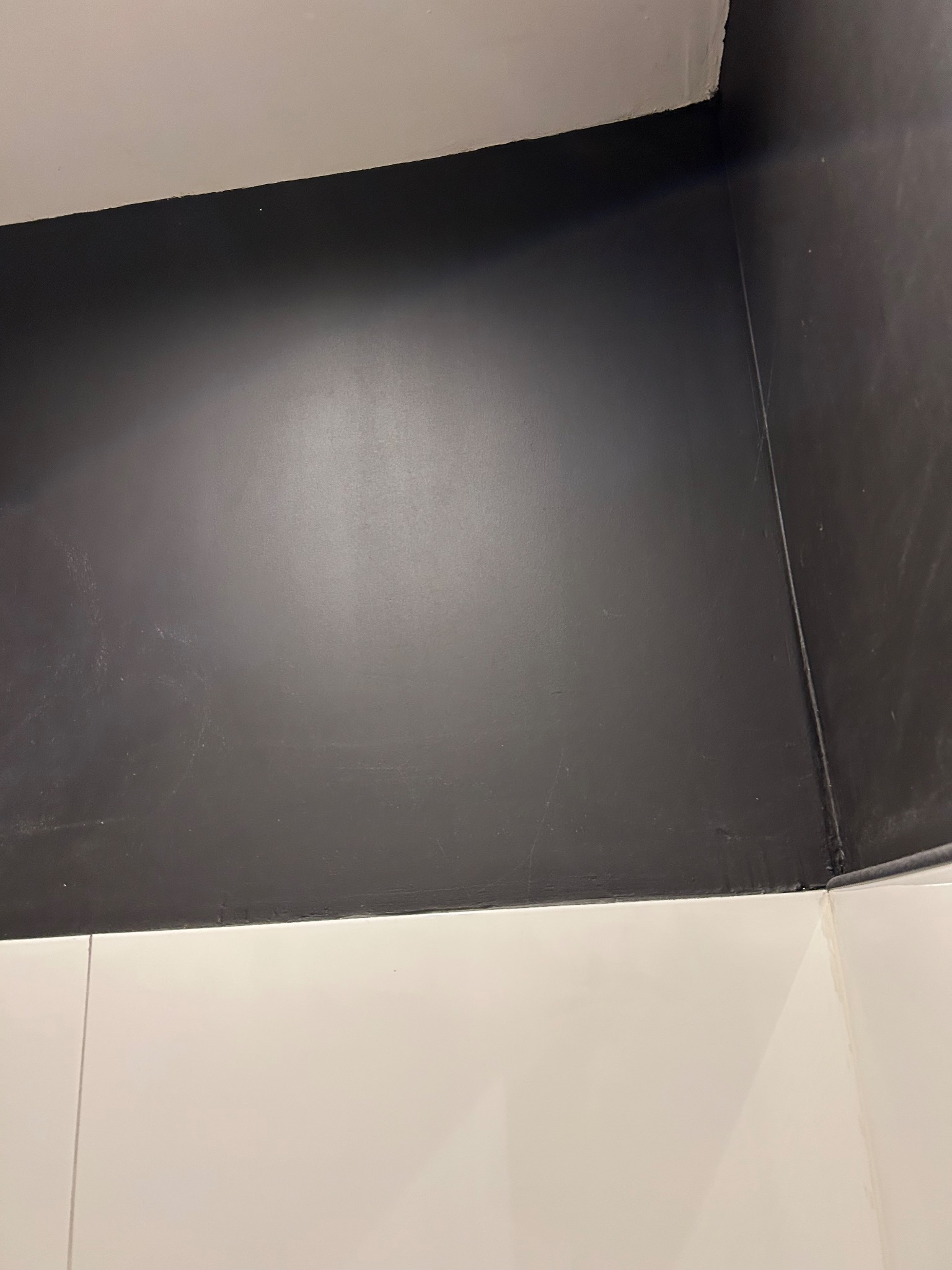 Ciemnoszara ściana nad białymi, błyszczącymi płytkami w rogu pomieszczenia. Widoczne łączenia płytek i niedoskonałości malowania.