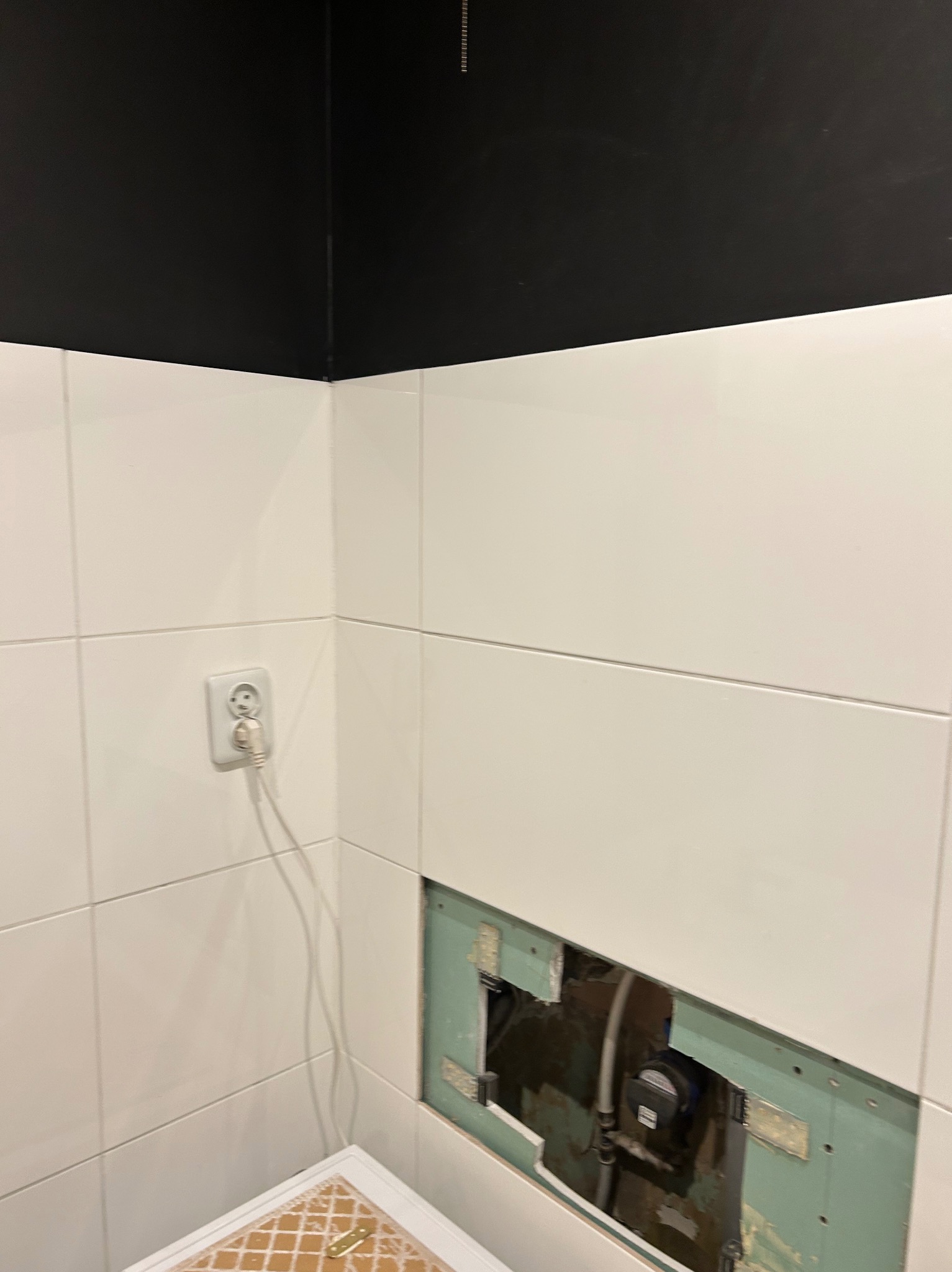 Białe płytki w łazience z czarnym sufitem, widoczne gniazdko i otwór w ścianie z instalacją. Etap prac wykończeniowych, widoczne elementy instalacji.
