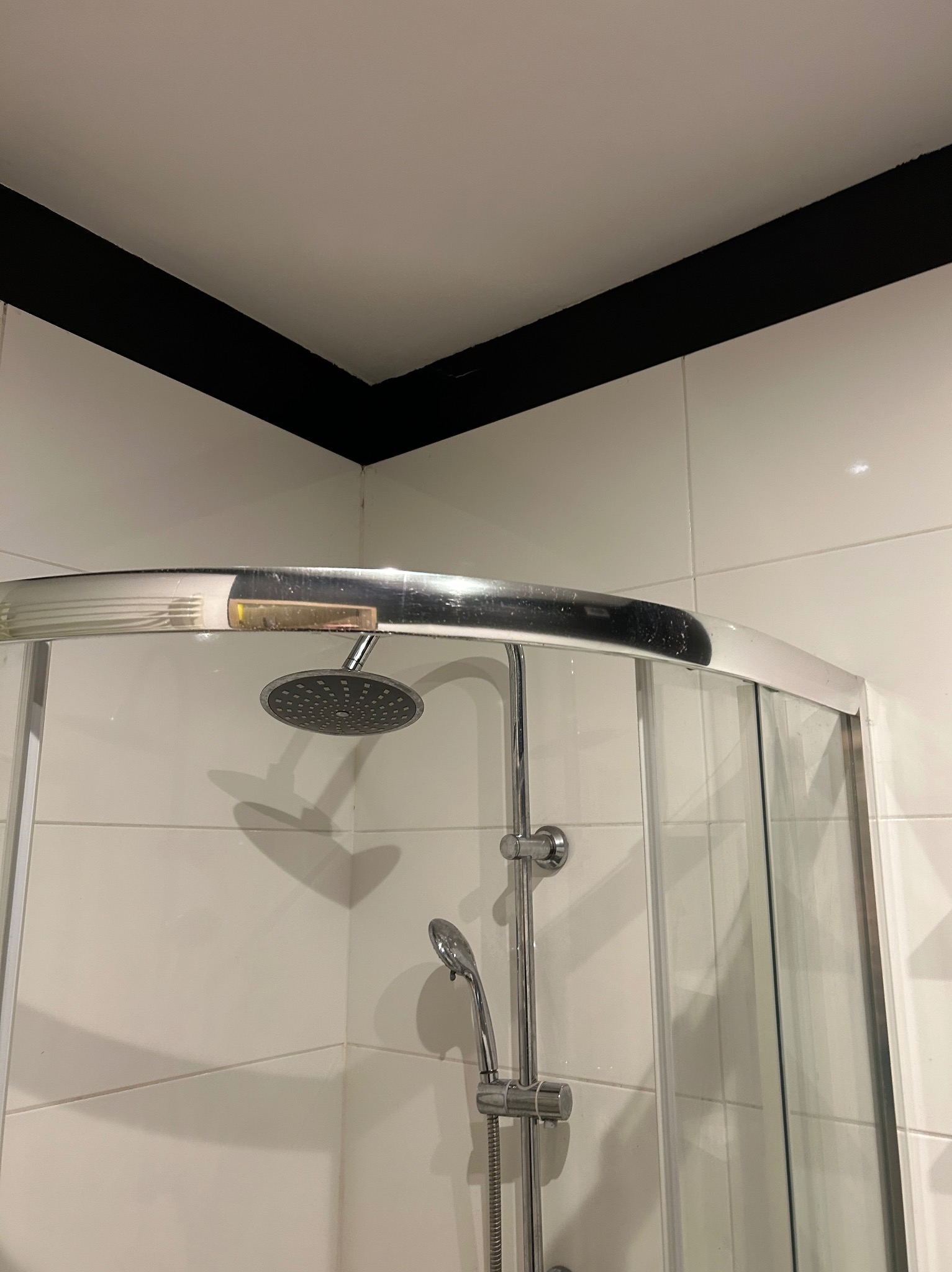 Kabina prysznicowa z chromowaną armaturą i białymi płytkami, widok z dołu, czarny pas sufitowy. Minimalistyczny design łazienki.