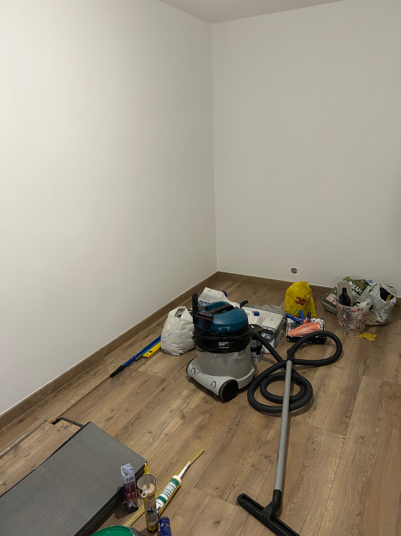 Pokój po remoncie z odkurzaczem przemysłowym, narzędziami i workami na podłodze z paneli. Widoczne świeżo pomalowane ściany i listwy przypodłogowe.