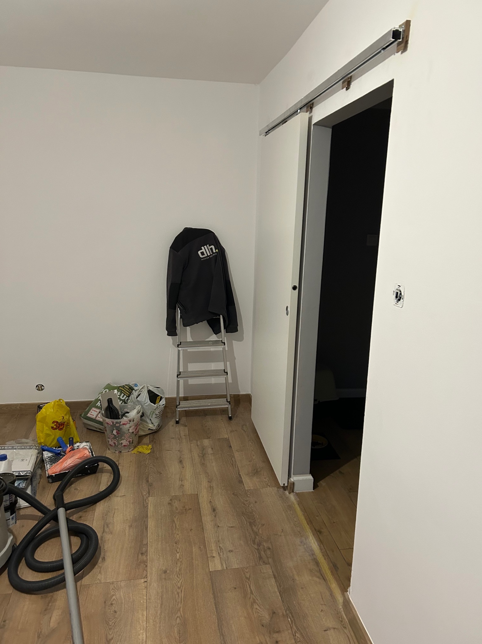 Wnętrze pokoju w trakcie montażu przesuwnych drzwi. Widoczna drabina, narzędzia i akcesoria malarskie. Podłoga wyłożona panelami imitującymi drewno.