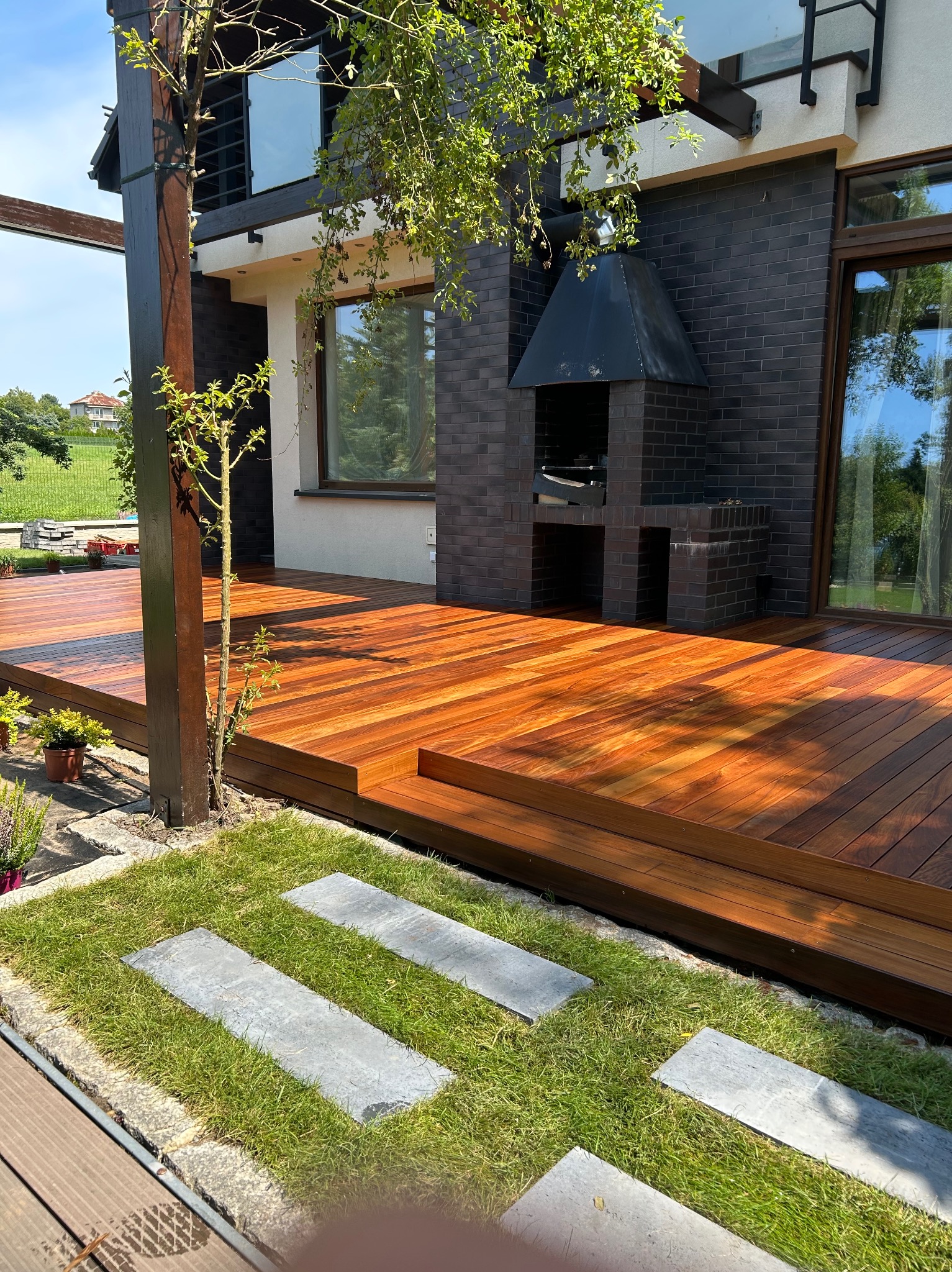 Nowoczesny taras z drewna egzotycznego, zintegrowany z grillem murowanym z ciemnej cegły. Kamienne płyty w trawie prowadzą do tarasu. Widok na dom i zieleń.