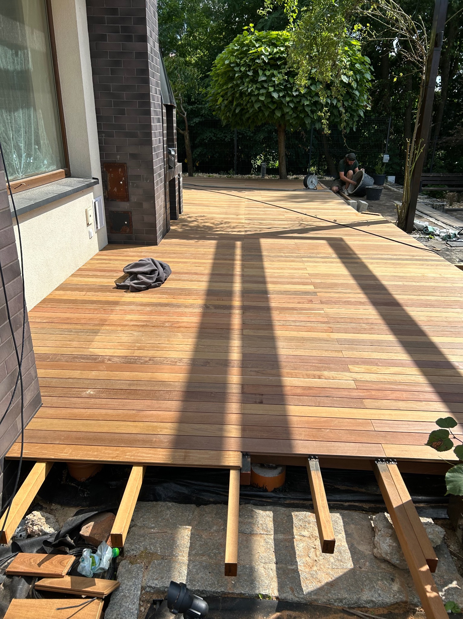 Nowa drewniana deska tarasowa w trakcie montażu, widoczne legary i podłoże. W tle osoba pracująca przy mieszaniu zaprawy. Słoneczny dzień, widok na ogród.