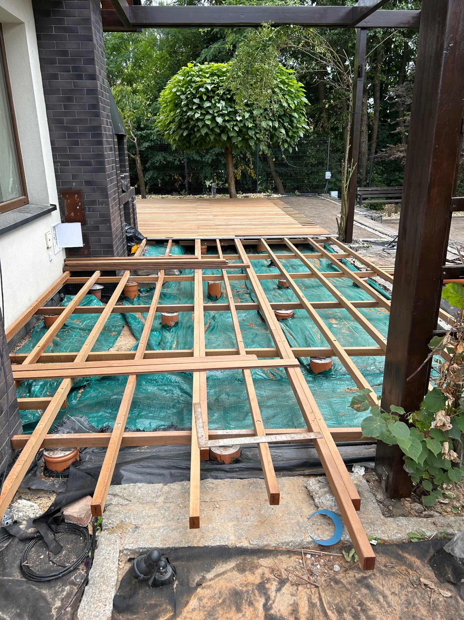 Drewniany szkielet tarasu w trakcie budowy, widoczne legary na zielonej folii, częściowo ułożona deska tarasowa, tło z zielenią. Ujęcie z góry, perspektywa szerokokątna.