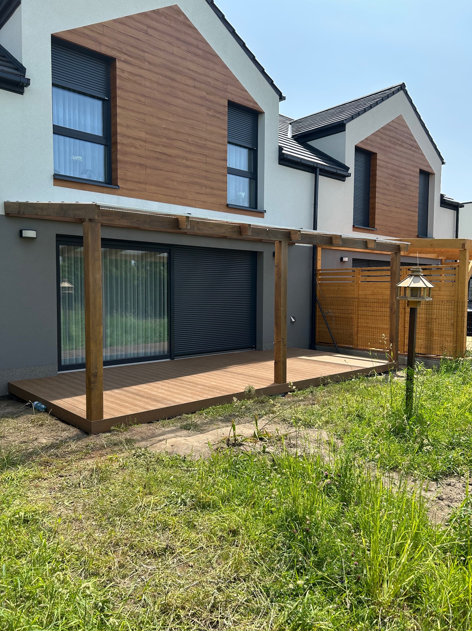 Nowoczesny taras z drewnianą podłogą i pergolą przy domu z drewnianą elewacją. Widoczne detale wykończenia i zadbany trawnik.