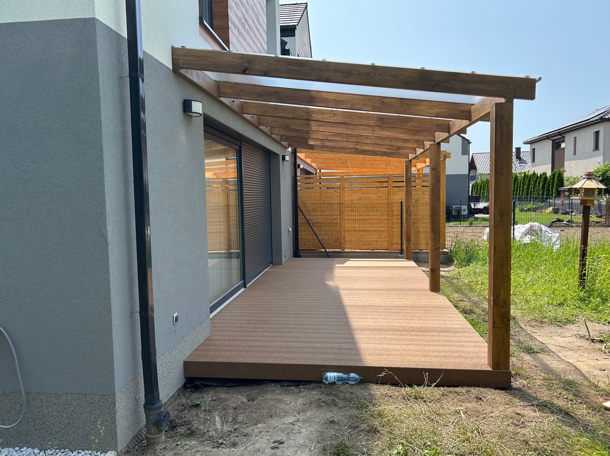 Zadaszony taras z drewnianą podłogą przy nowoczesnym domu. Widoczne elementy konstrukcyjne z drewna i przesuwne drzwi balkonowe.