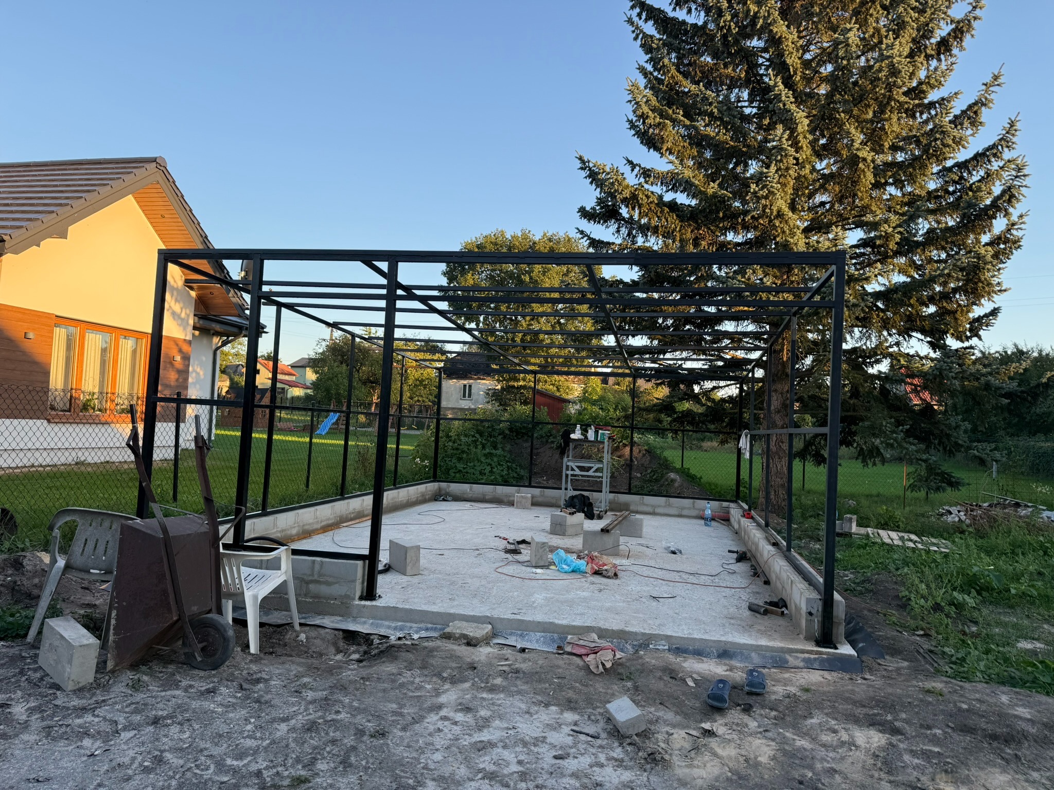 Stalowa konstrukcja szkieletowa w ogrodzie, prawdopodobnie pod przyszłą pergolę. Widoczne fundamenty i narzędzia, co sugeruje etap budowy. Ujęcie w słoneczny dzień.