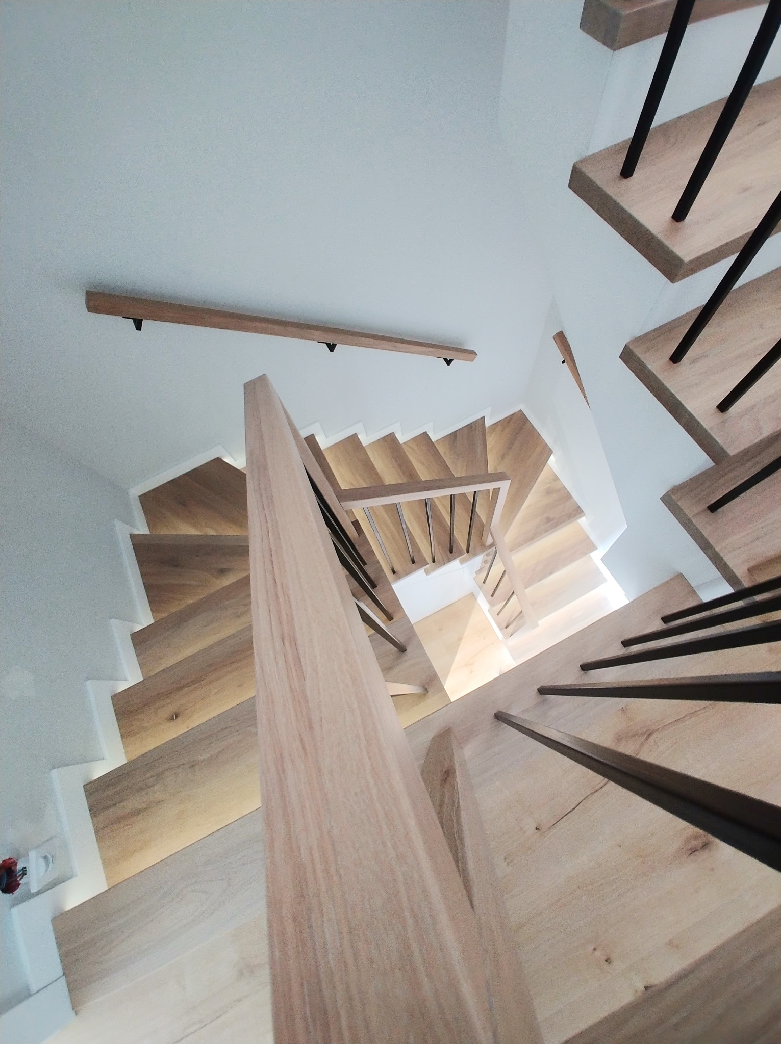 Spiralne schody z jasnego drewna z czarnymi, metalowymi balustradami, oświetlone od spodu, widziane z góry pod nietypowym kątem, ukazujące nowoczesny design.