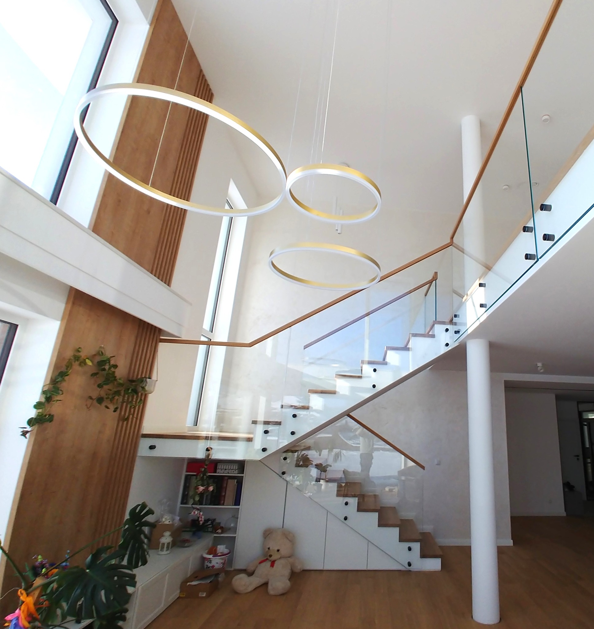 Nowoczesne schody z drewnianymi stopniami i szklaną balustradą, oświetlone wiszącymi lampami w kształcie okręgów. W tle drewniana ściana i duże okno.