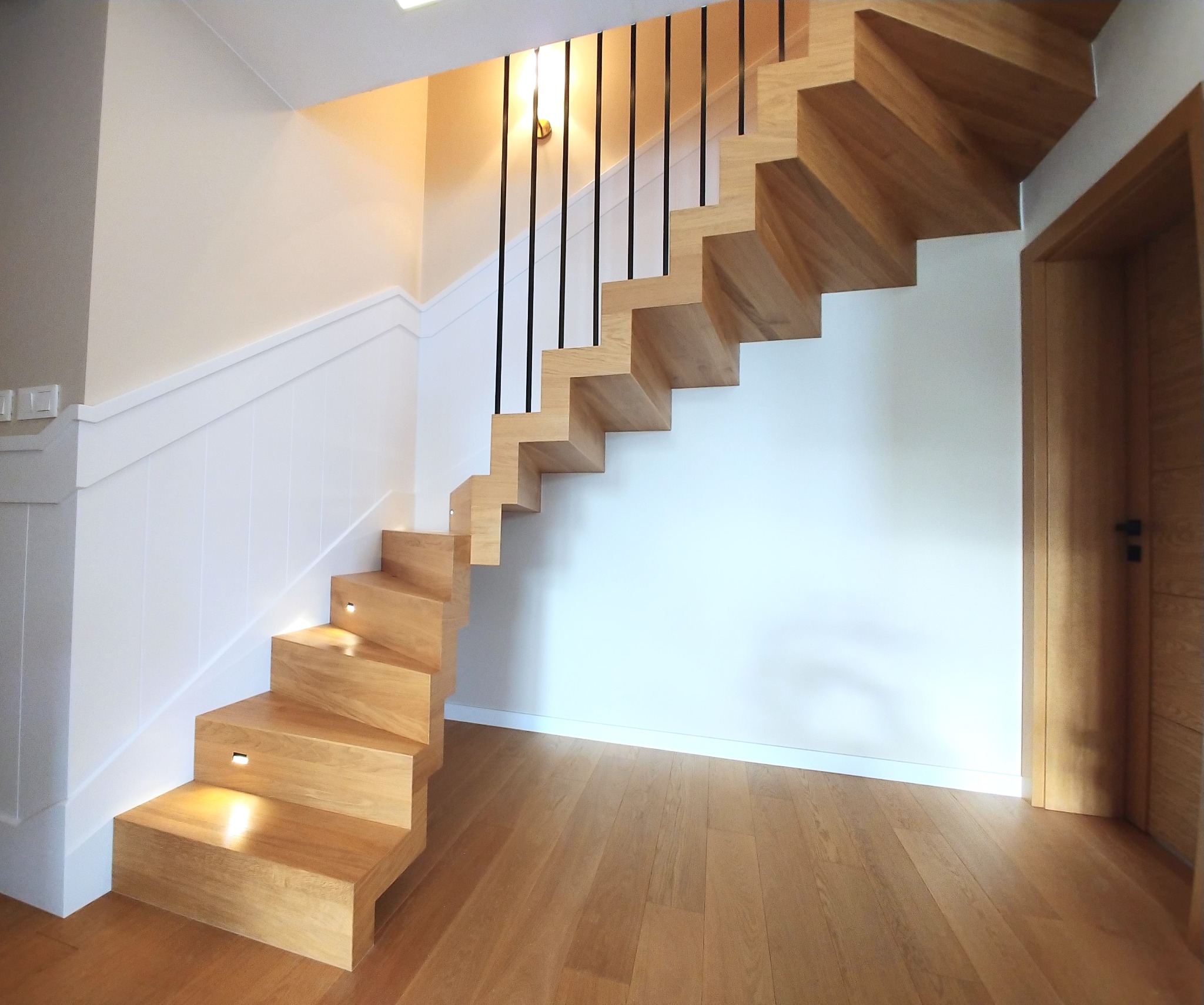 Nowoczesne, drewniane schody wspornikowe z czarną balustradą i subtelnym oświetleniem LED w stopniach, na tle jasnej ściany i drewnianej podłogi.
