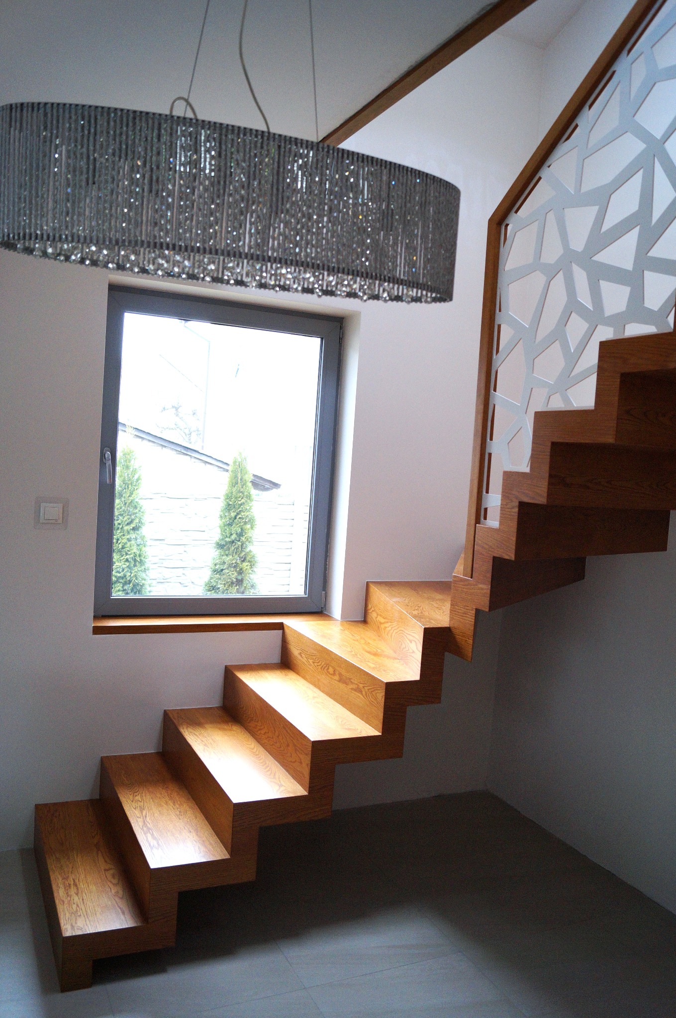 Drewniane schody wspornikowe z ażurową balustradą, oświetlone dekoracyjnym żyrandolem, umieszczone przy oknie z widokiem na zieleń. Nowoczesny design.