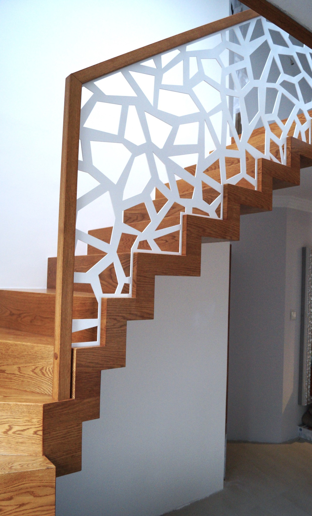 Nowoczesne schody z drewnianymi stopniami i białą, ażurową balustradą o geometrycznym wzorze. Jasne wnętrze, minimalistyczny design.