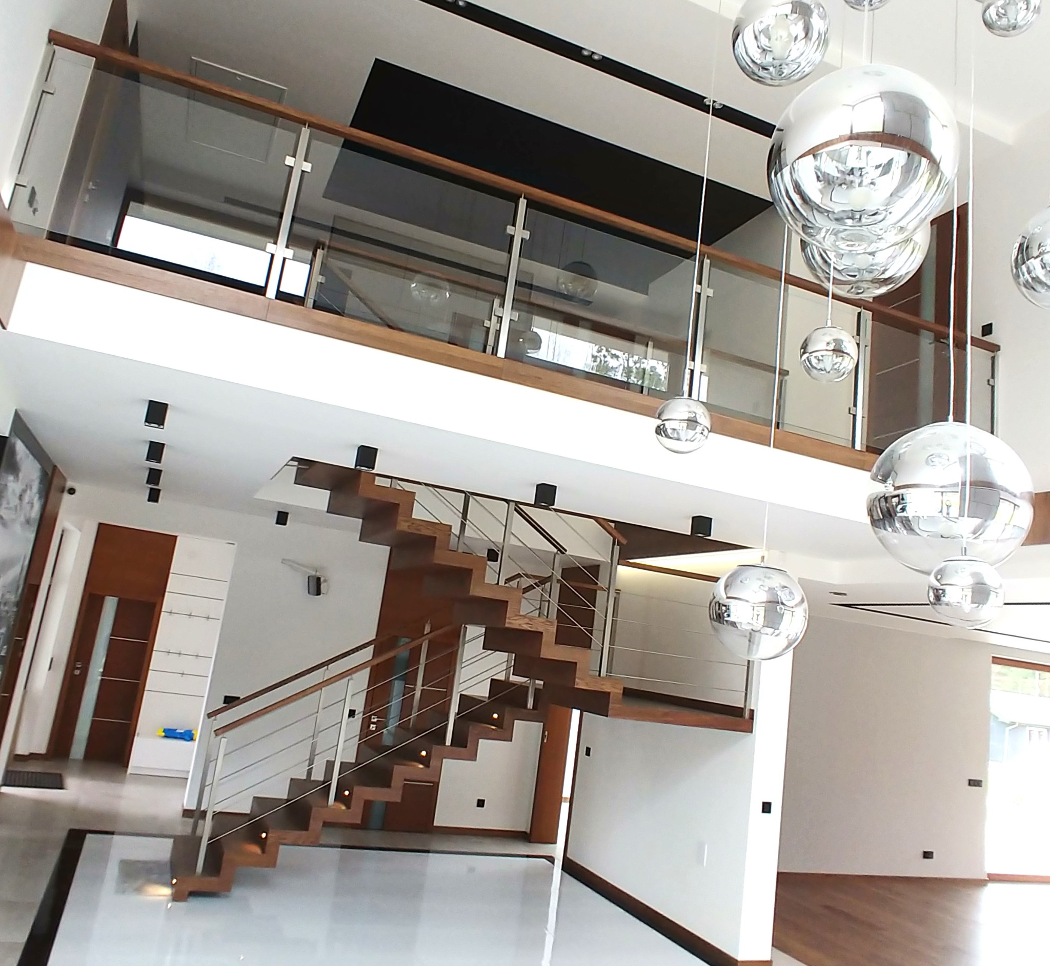 Nowoczesne schody drewniane z balustradą szklaną i metalowymi elementami w przestronnym wnętrzu z designerskimi lampami. Widok z dołu, podkreślający architekturę.