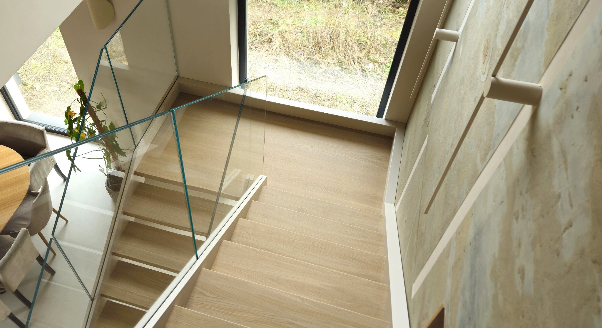 Jasne, drewniane schody z przezroczystą szklaną balustradą, widziane z góry. Obok schodów stół z krzesłami i roślina doniczkowa. Duże okno w tle.