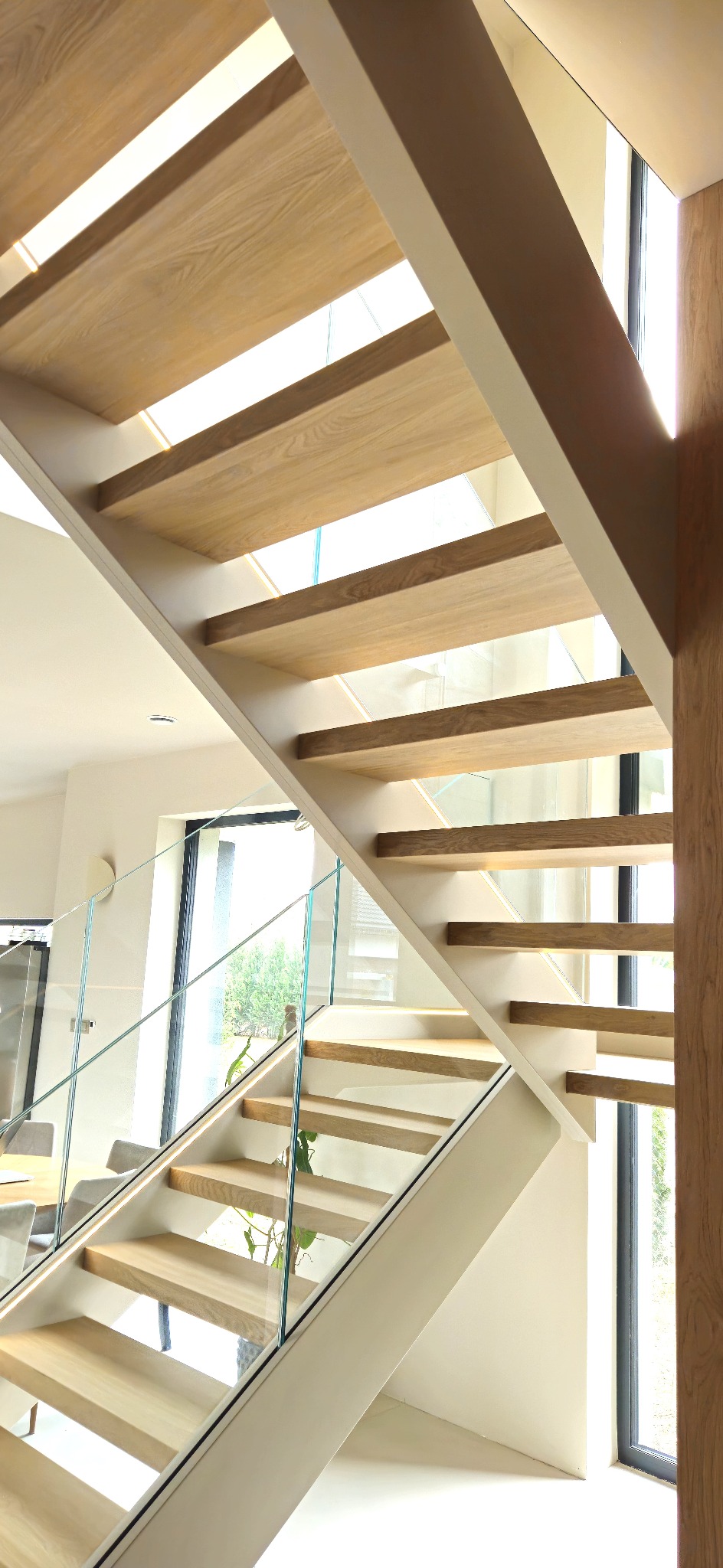 Nowoczesne schody z jasnego drewna z przezroczystą szklaną balustradą, oświetlone od spodu, w minimalistycznym wnętrzu z dużym oknem i rośliną doniczkową.