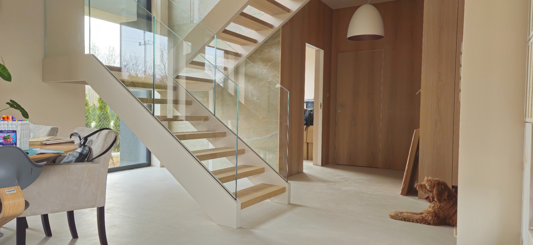 Nowoczesne schody z drewnianymi stopniami i szklaną balustradą w jasnym wnętrzu domu w Dobre. Widoczny pies leżący na podłodze.