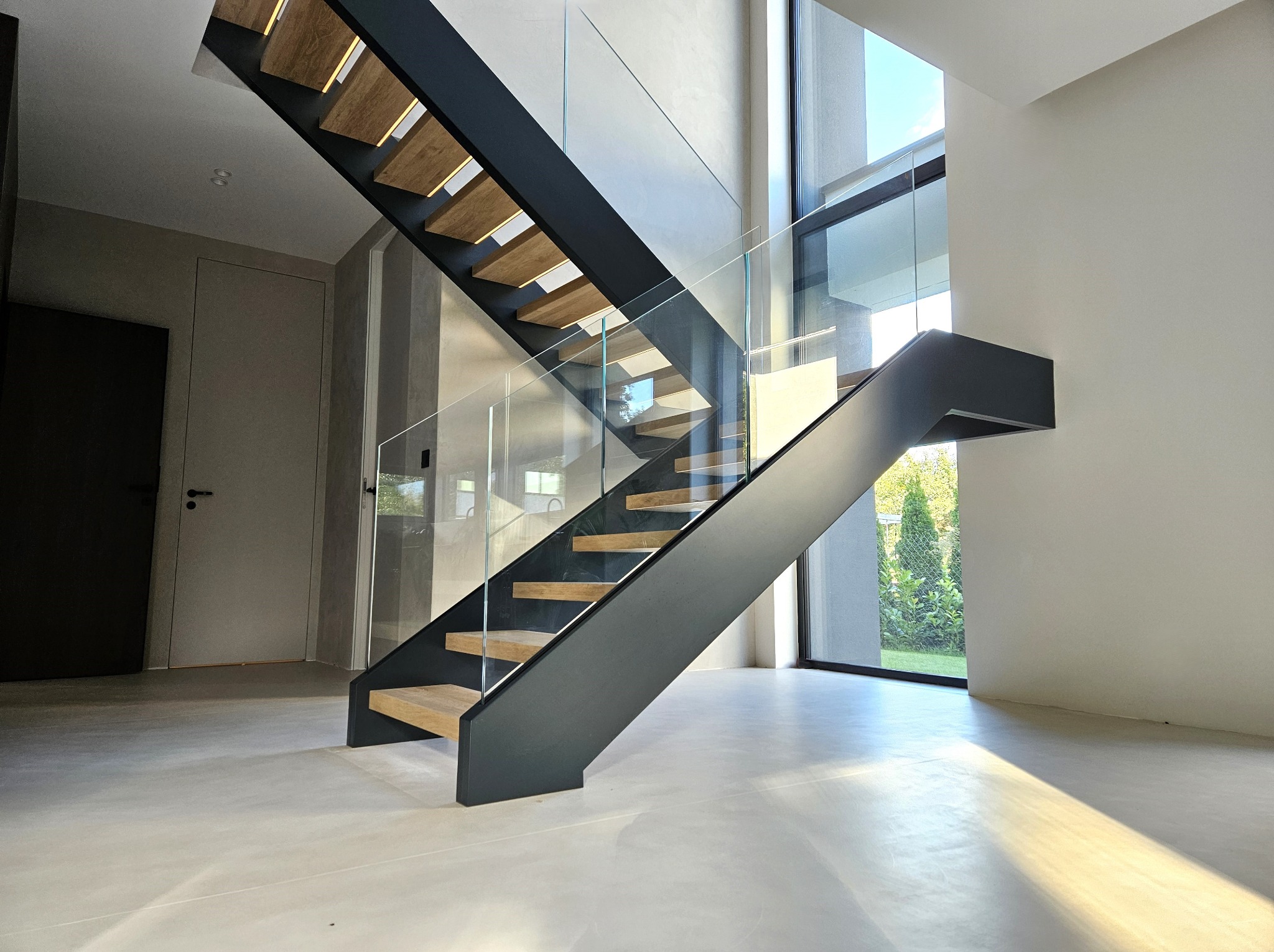 Nowoczesne schody wspornikowe z drewnianymi stopniami i szklaną balustradą w jasnym wnętrzu z dużym oknem. Minimalistyczny design, gra światła na podłodze.