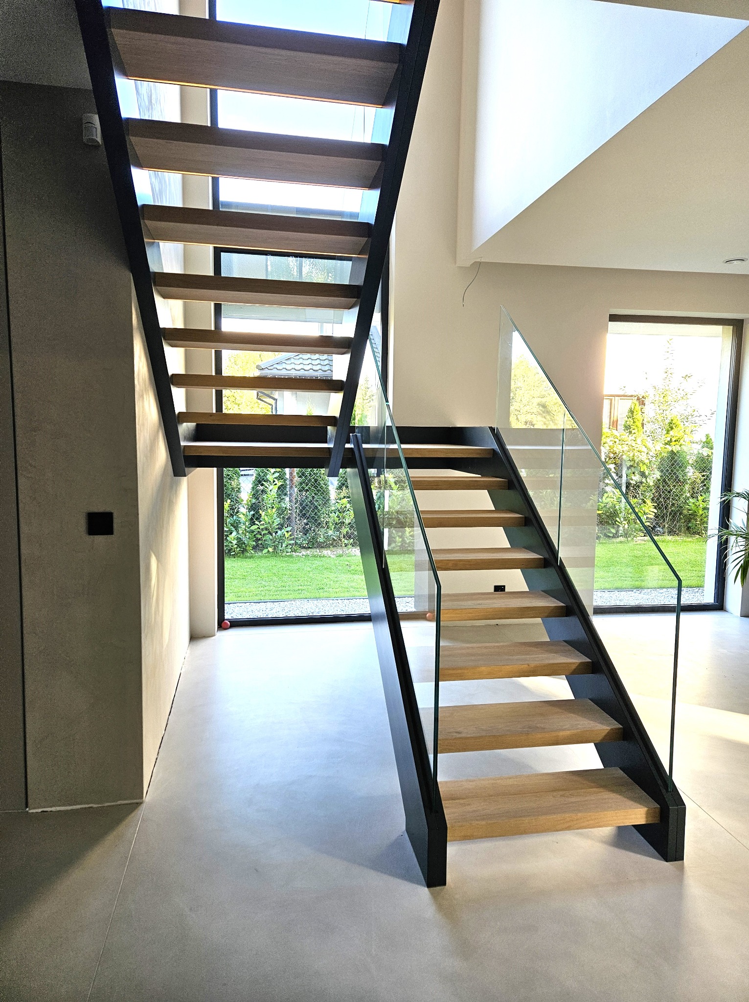 Nowoczesne schody z drewnianymi stopniami i szklaną balustradą, czarna konstrukcja, widok na ogród przez okno, minimalistyczny design, jasne wnętrze.