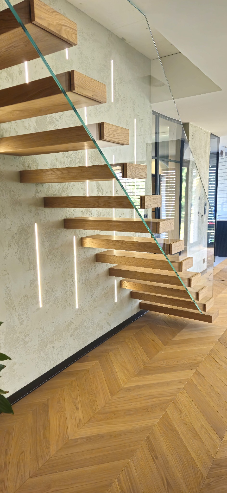 Nowoczesne schody wspornikowe z drewna dębowego, zintegrowane z szklaną balustradą i oświetleniem LED, na tle ściany o fakturze betonu i parkietu w jodełkę.