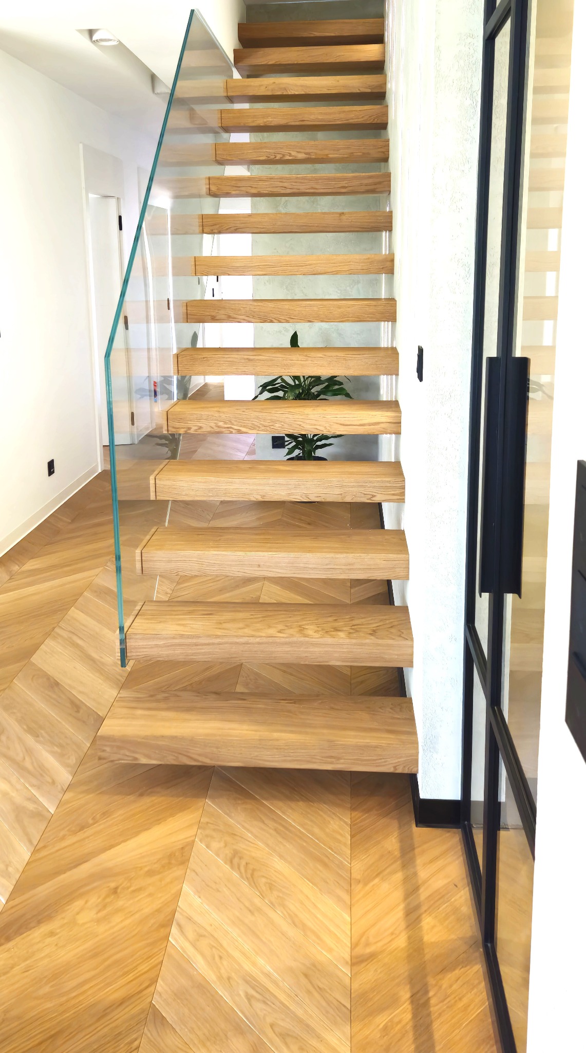 Nowoczesne schody z drewnianymi stopniami i szklaną balustradą, oświetlone naturalnym światłem, z parkietem ułożonym w jodełkę na podłodze.