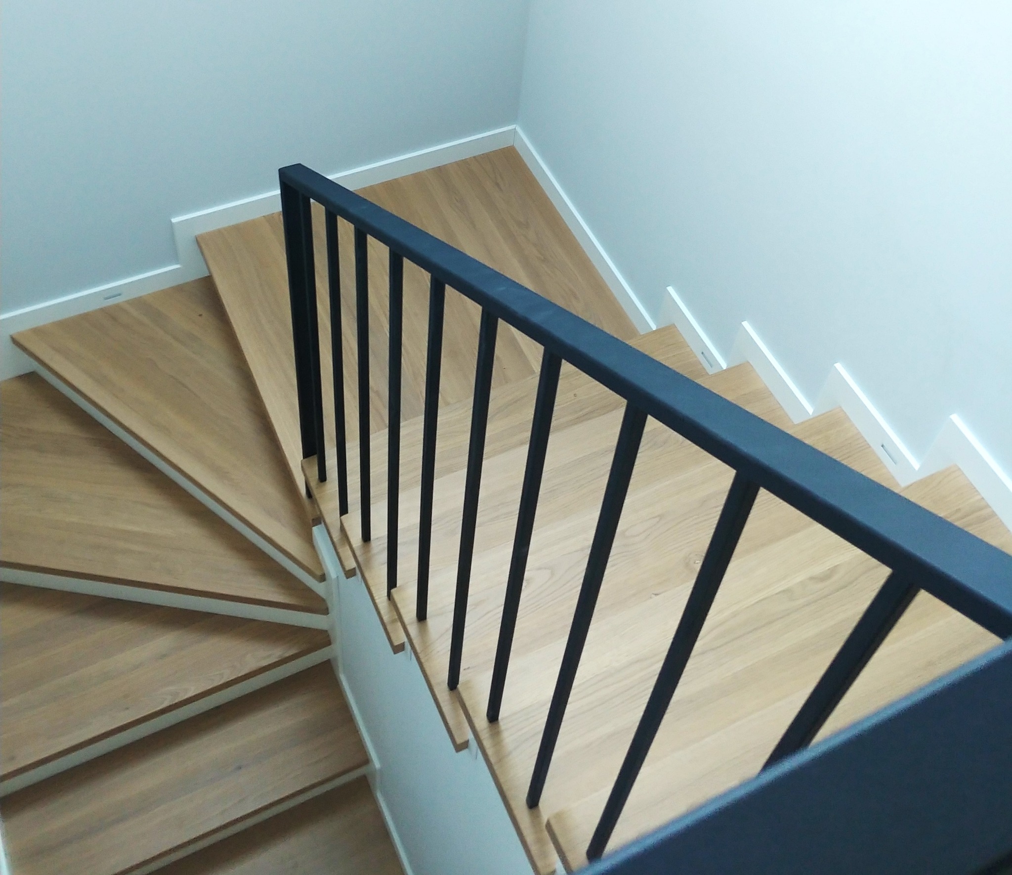 Drewniane schody z czarną, metalową balustradą, widok z góry. Jasne drewno stopni kontrastuje z ciemną balustradą. Minimalistyczny design, nowoczesne wykończenie.