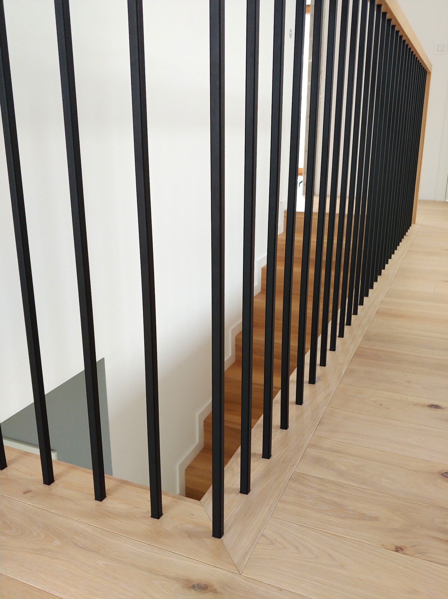 Minimalistyczna balustrada z czarnymi, pionowymi słupkami na jasnej drewnianej podłodze i schodach. Nowoczesny design, proste linie, neutralna kolorystyka.