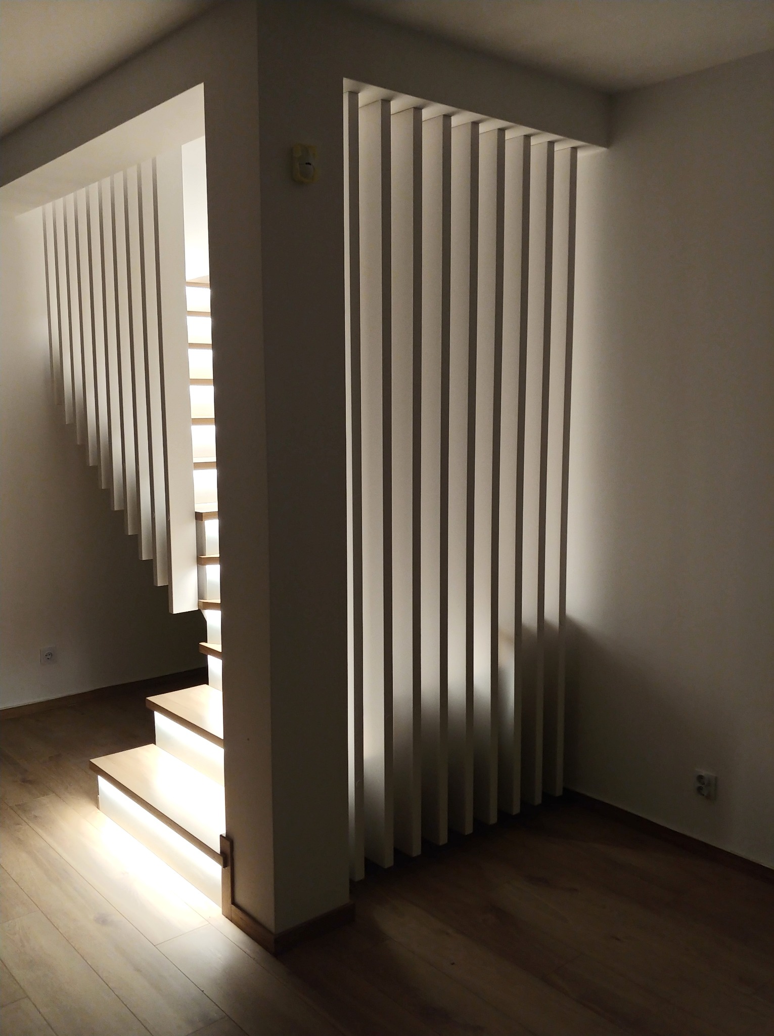 Nowoczesne schody z podświetleniem LED i ażurową balustradą z pionowych listew, tworzące minimalistyczny design w jasnym wnętrzu. Widoczne drewniane stopnie i podłoga.