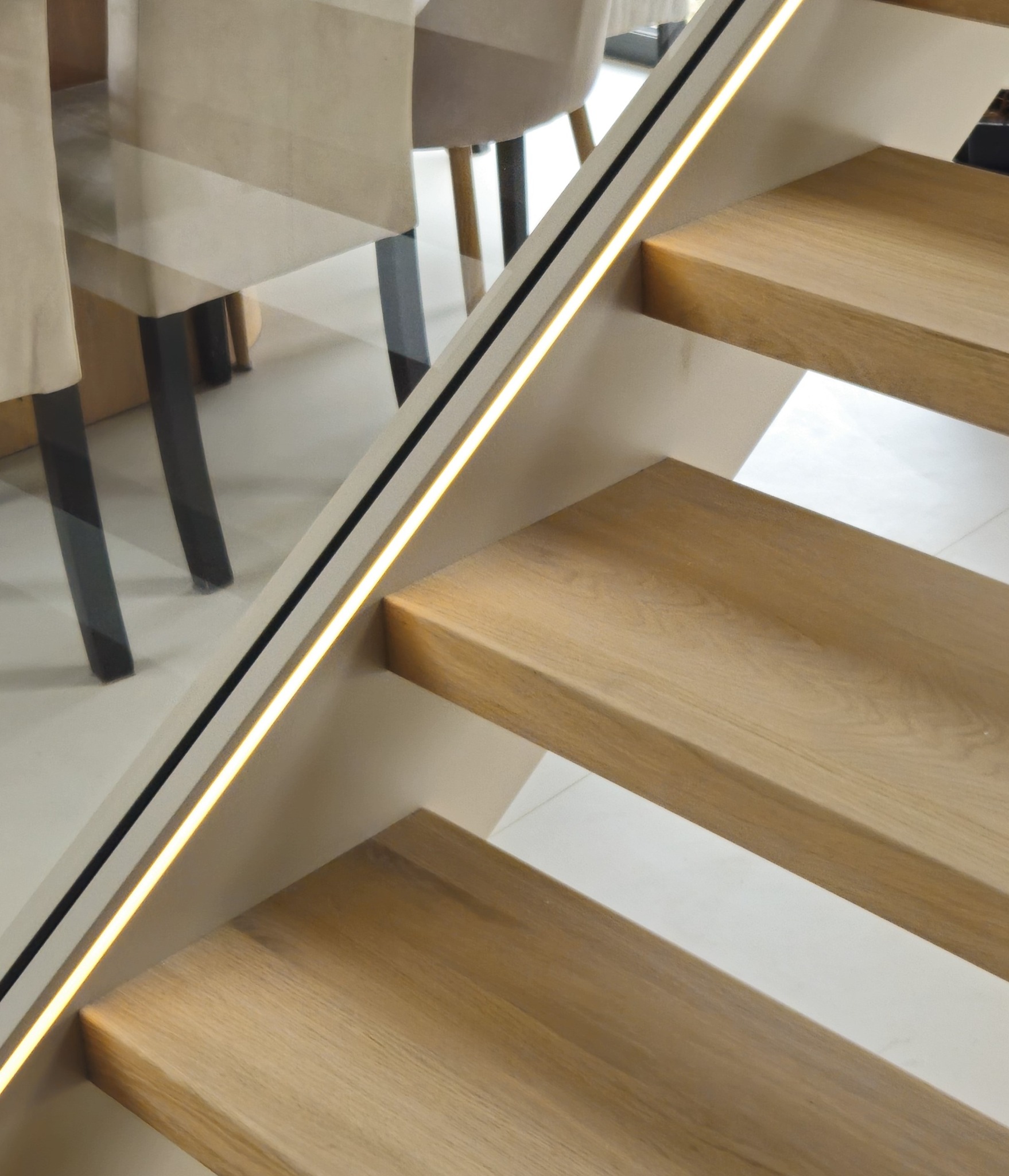 Nowoczesne schody z drewna dębowego z podświetlaną balustradą szklaną. Widoczne trzy stopnie i fragment krzesła w tle. Minimalistyczny design.