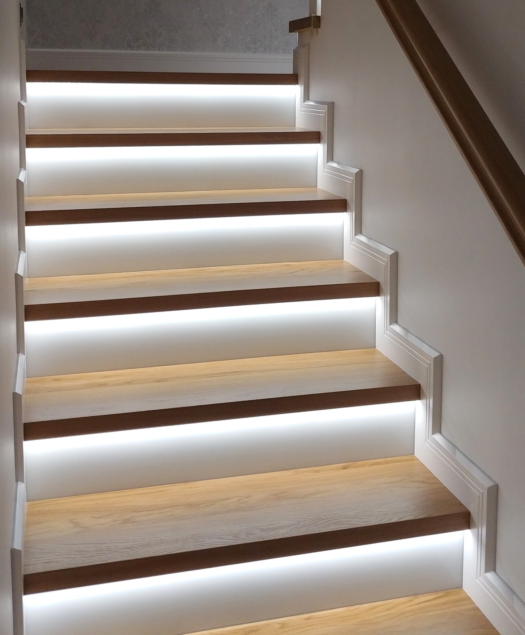 Nowoczesne schody z podświetleniem LED, drewniane stopnie o ciepłym odcieniu, białe boki i listwy przypodłogowe. Minimalistyczny design, subtelne oświetlenie.