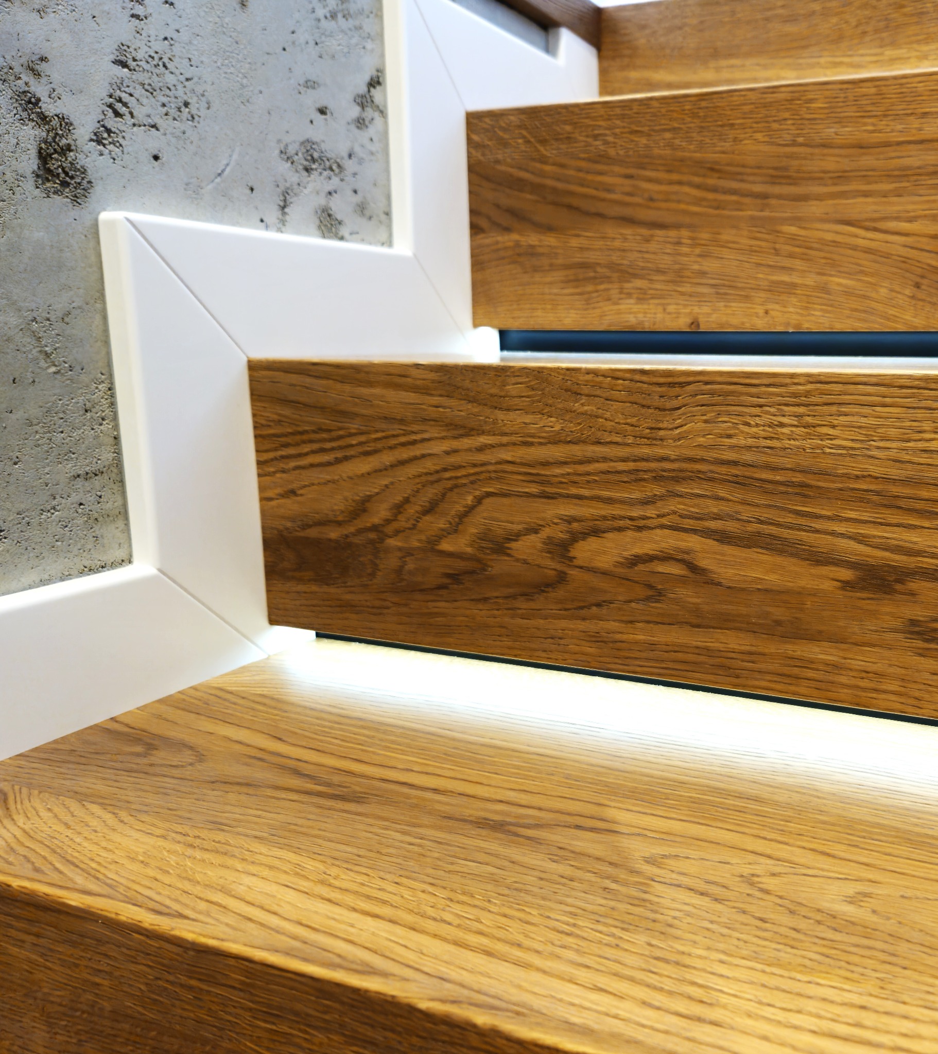 Detale drewnianych schodów z białymi listwami i subtelnym oświetleniem LED, ukazujące precyzję wykonania i nowoczesny design. Widoczna faktura drewna i surowy beton na ścianie.