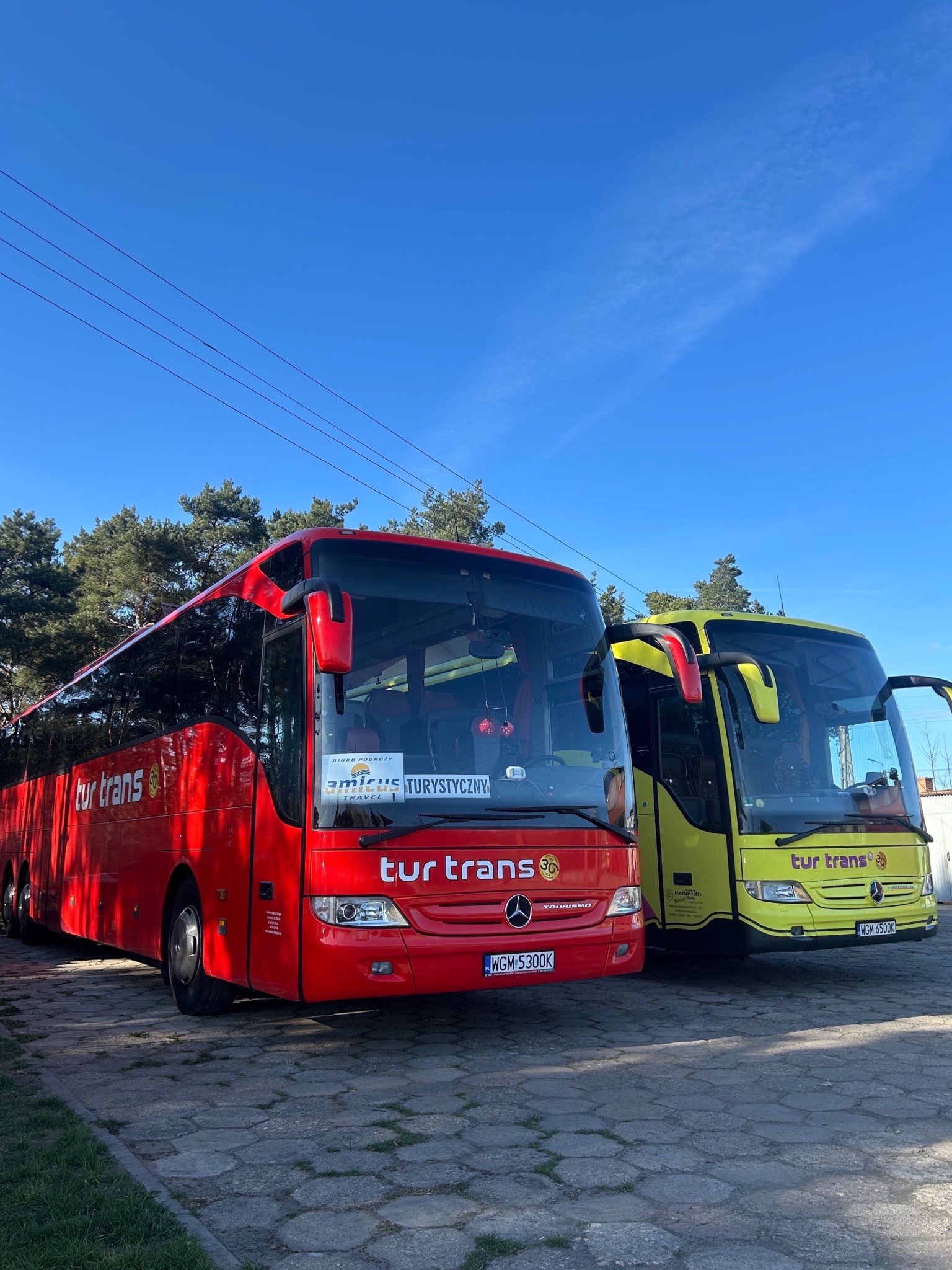 Dwa autokary Tur Trans: czerwony i żółty, stojące obok siebie na brukowanym placu, pod błękitnym niebem. Widoczne logo firmy i numery rejestracyjne.