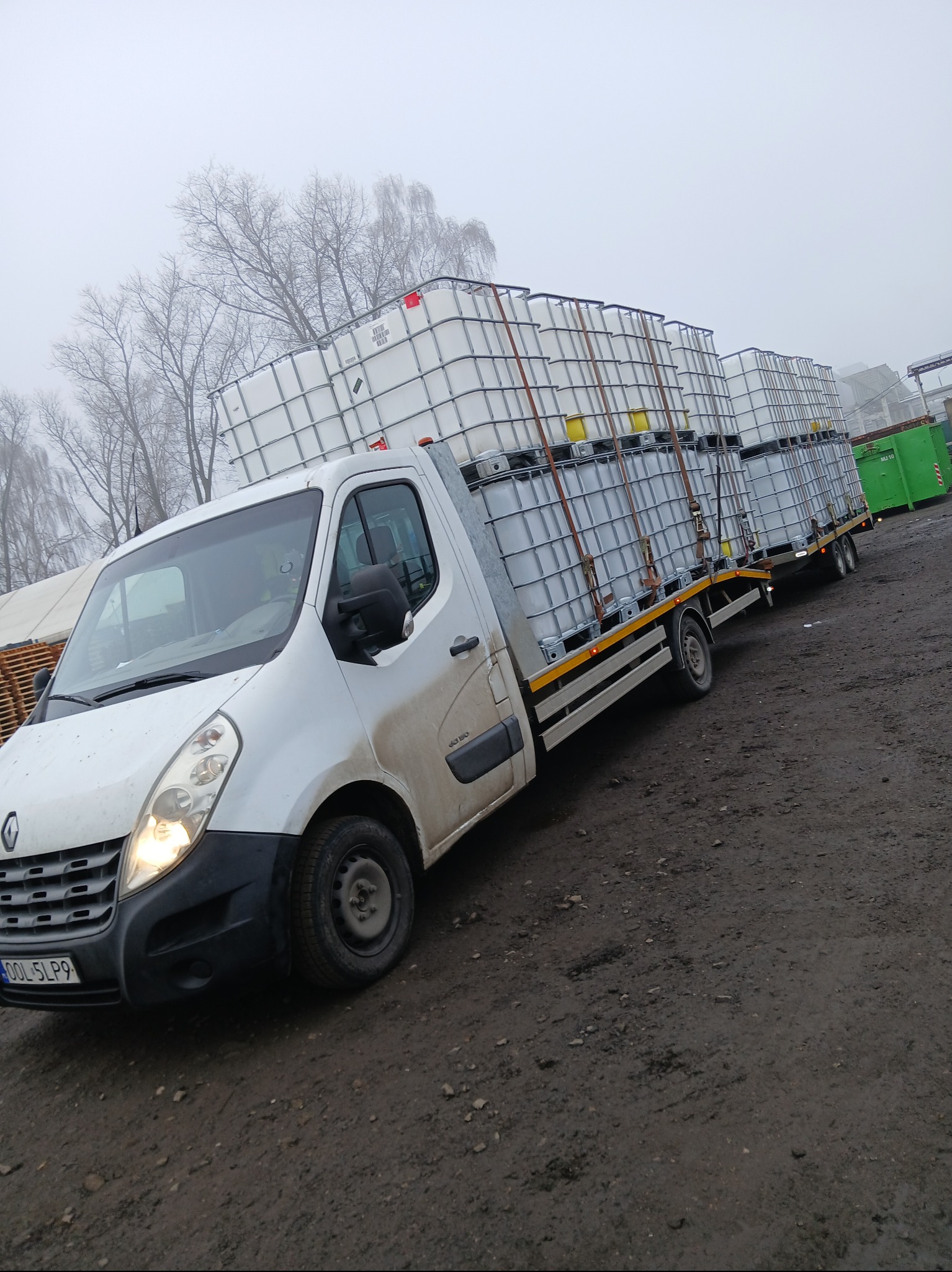 Biały Renault Master z przyczepą transportuje zbiorniki IBC na błotnistym terenie. Widoczne drzewa i mgła w tle. Tablica rejestracyjna OOL 5LP9.