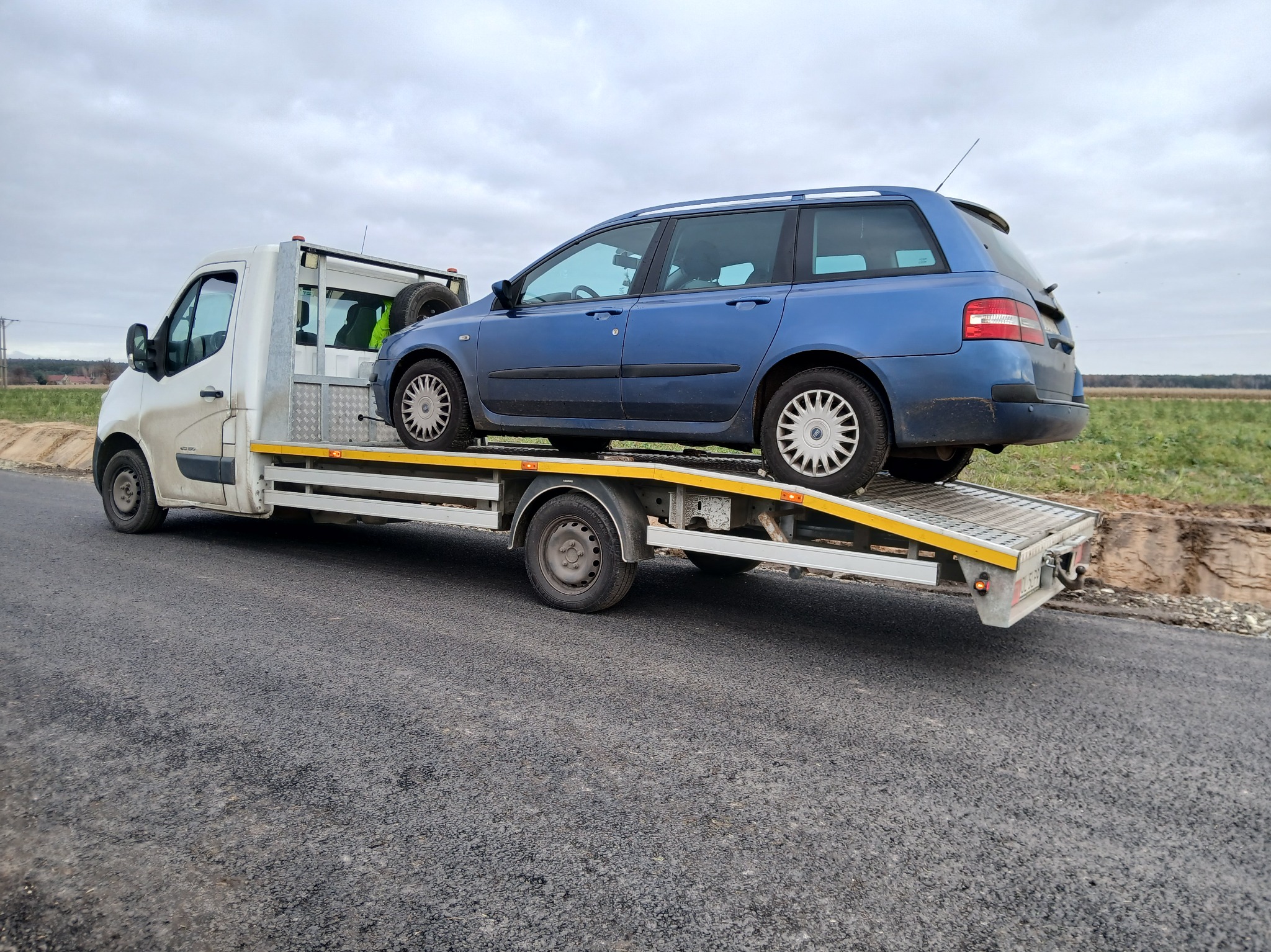 Błękitne kombi na białej platformie pomocy drogowej, widok z boku na szarej drodze, w tle pole i pochmurne niebo. Koło zapasowe za kabiną.