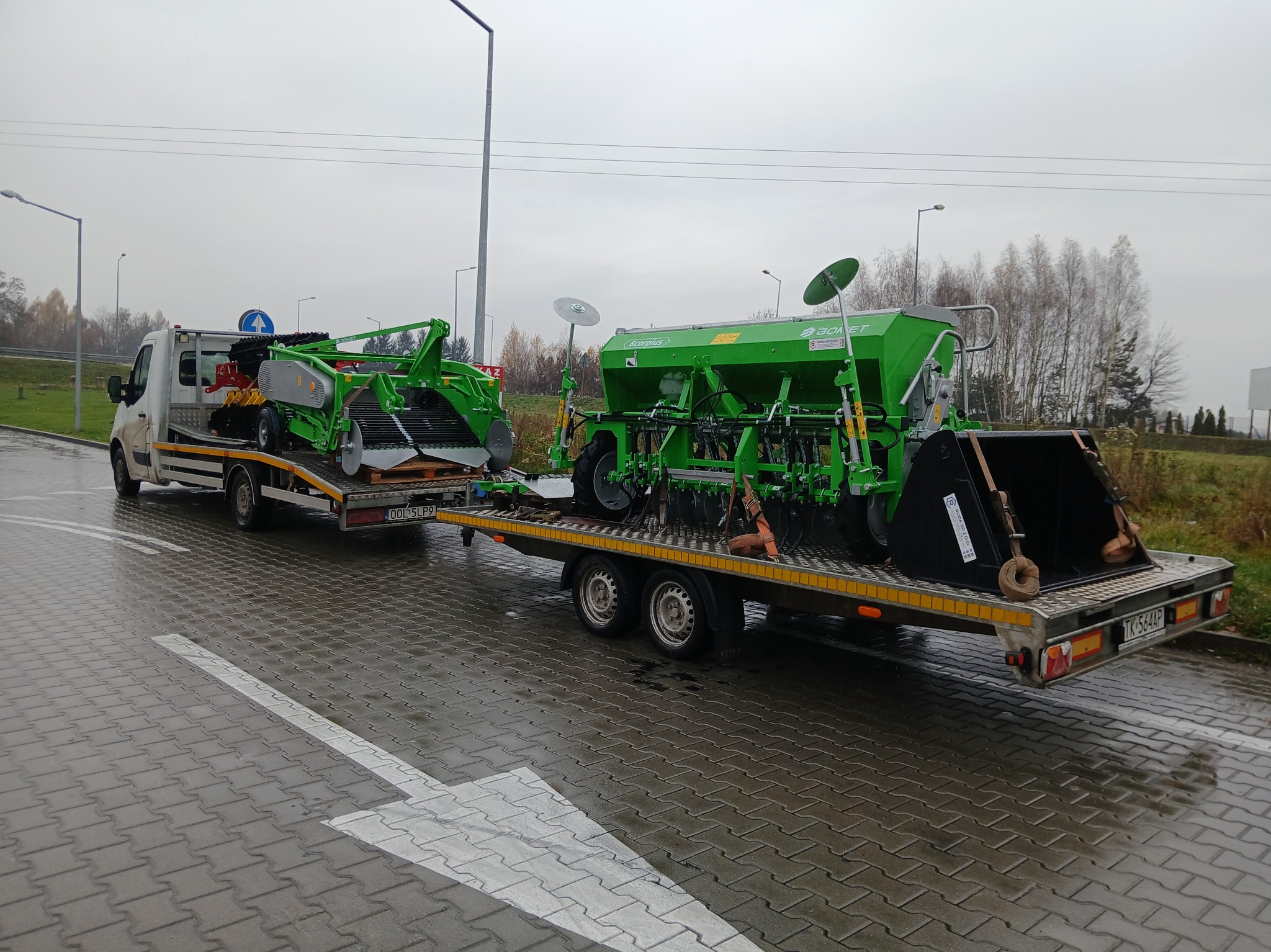 Transport dwóch zielonych maszyn rolniczych na niskopodwoziowej przyczepie, ciągniętej przez biały samochód dostawczy na mokrej nawierzchni.