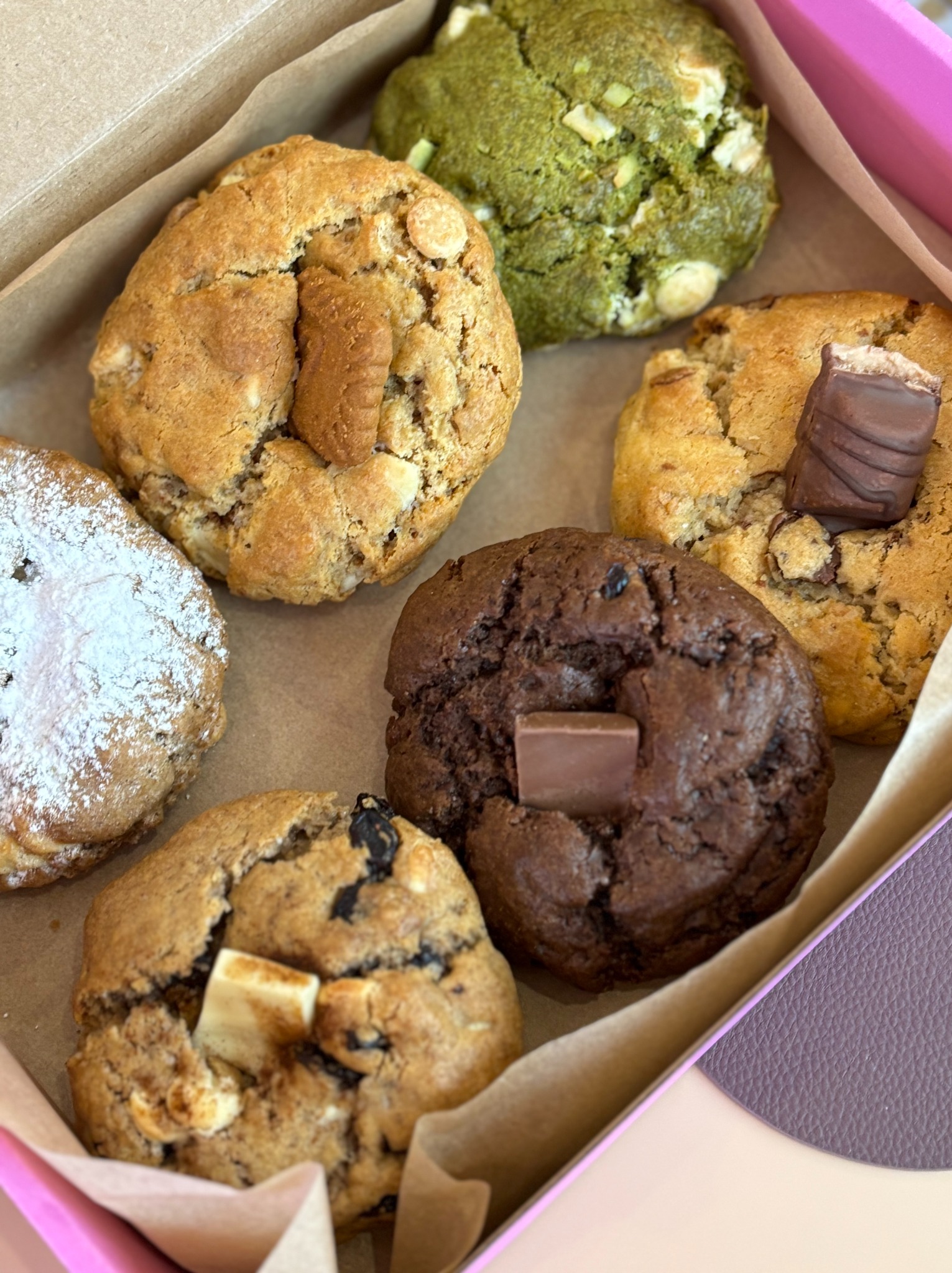 Różnorodne, ręcznie robione ciasteczka w kartonowym pudełku z różową obwódką. Wśród nich: czekoladowe, matcha i posypane cukrem pudrem.