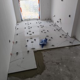 R-BUD - Układanie białych płytek imitujących marmur na podłodze w jasnym pomieszczeniu. Widoczne klipsy poziomujące i paczka z akcesoriami.