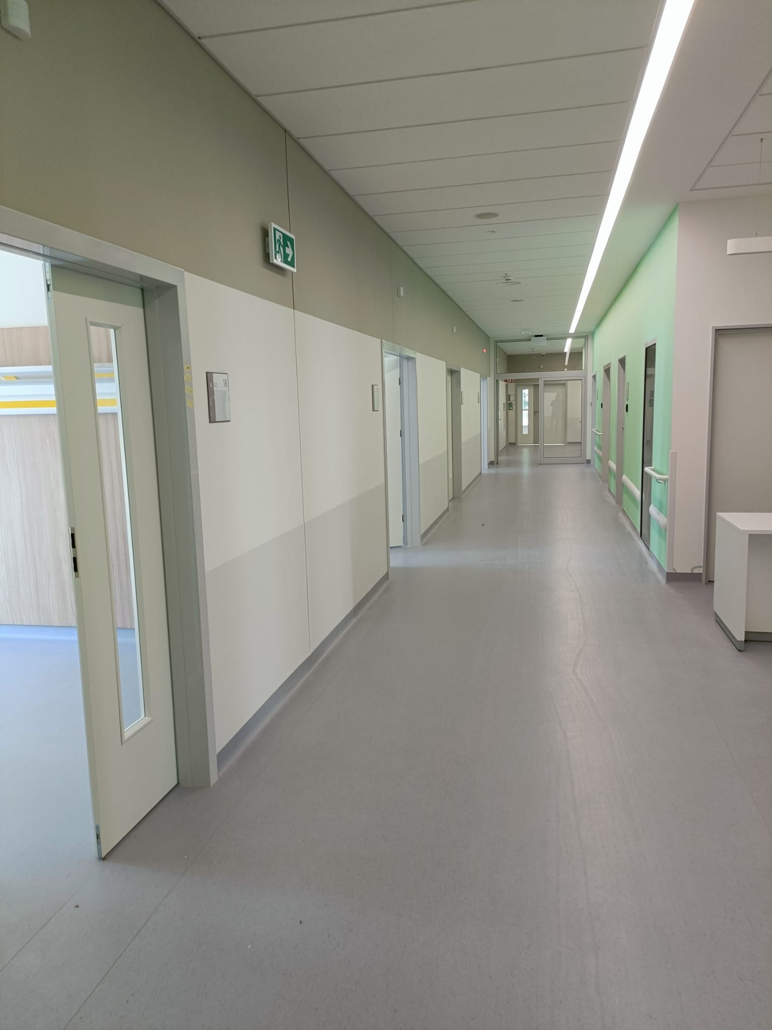Długi korytarz w budynku użyteczności publicznej z szarymi panelami ściennymi i podłogą, oświetlony liniowymi lampami sufitowymi i znakami ewakuacyjnymi.