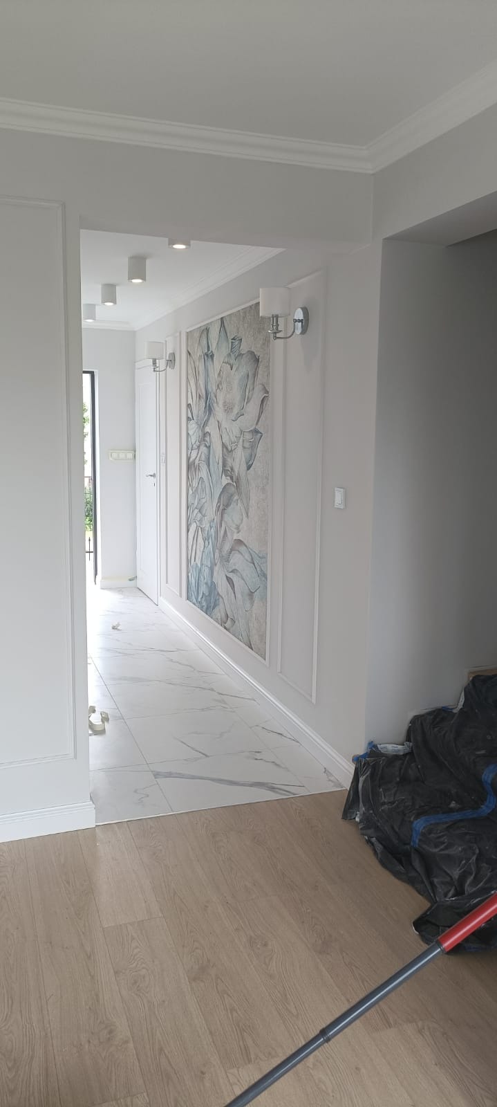 Wykończony korytarz w jasnych barwach z panelami podłogowymi i marmurową posadzką. Na ścianie dekoracyjny panel z motywem roślinnym i kinkietami. Listwy przypodłogowe.