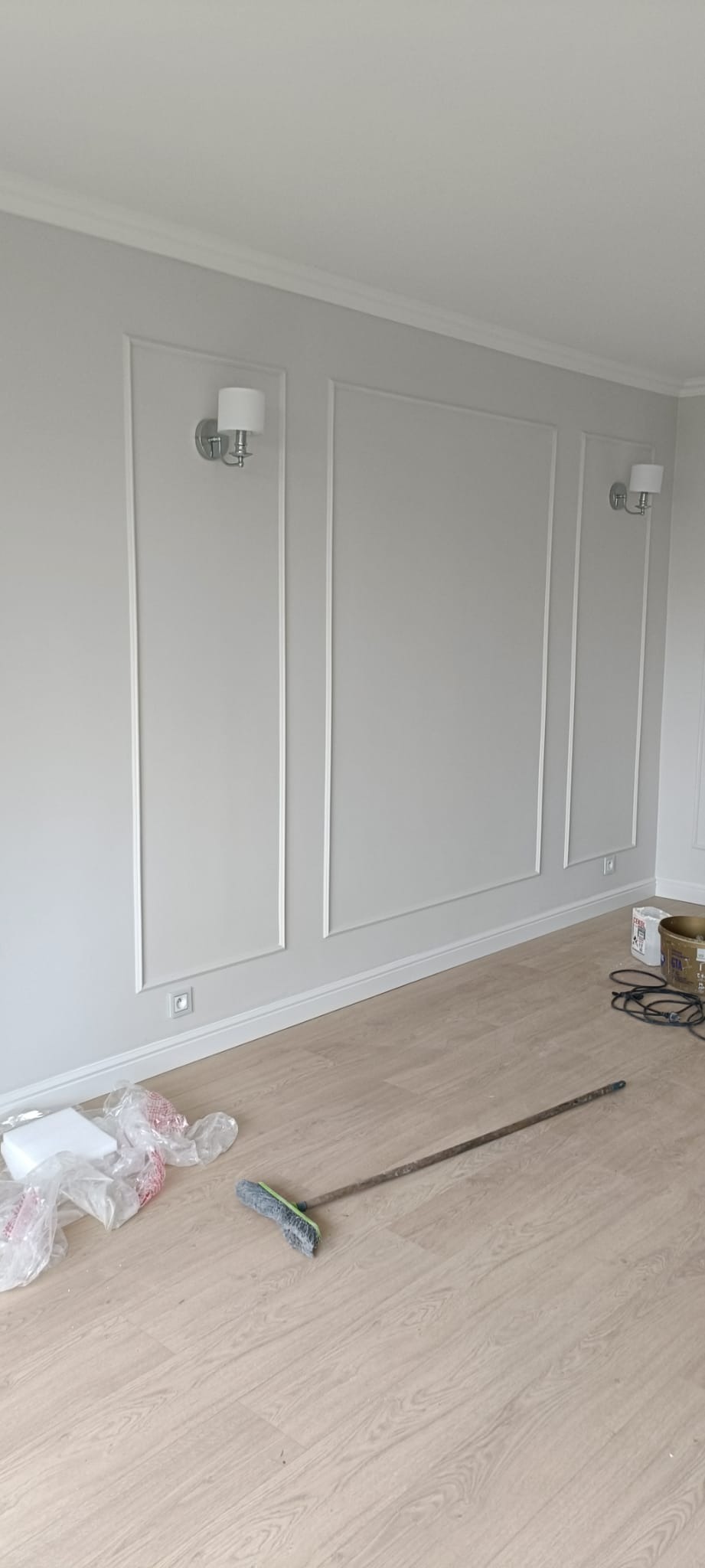 Jasnoszary pokój po remoncie: listwy dekoracyjne na ścianach, lampy ścienne, panele podłogowe. Widoczne akcesoria malarskie i szczotka.