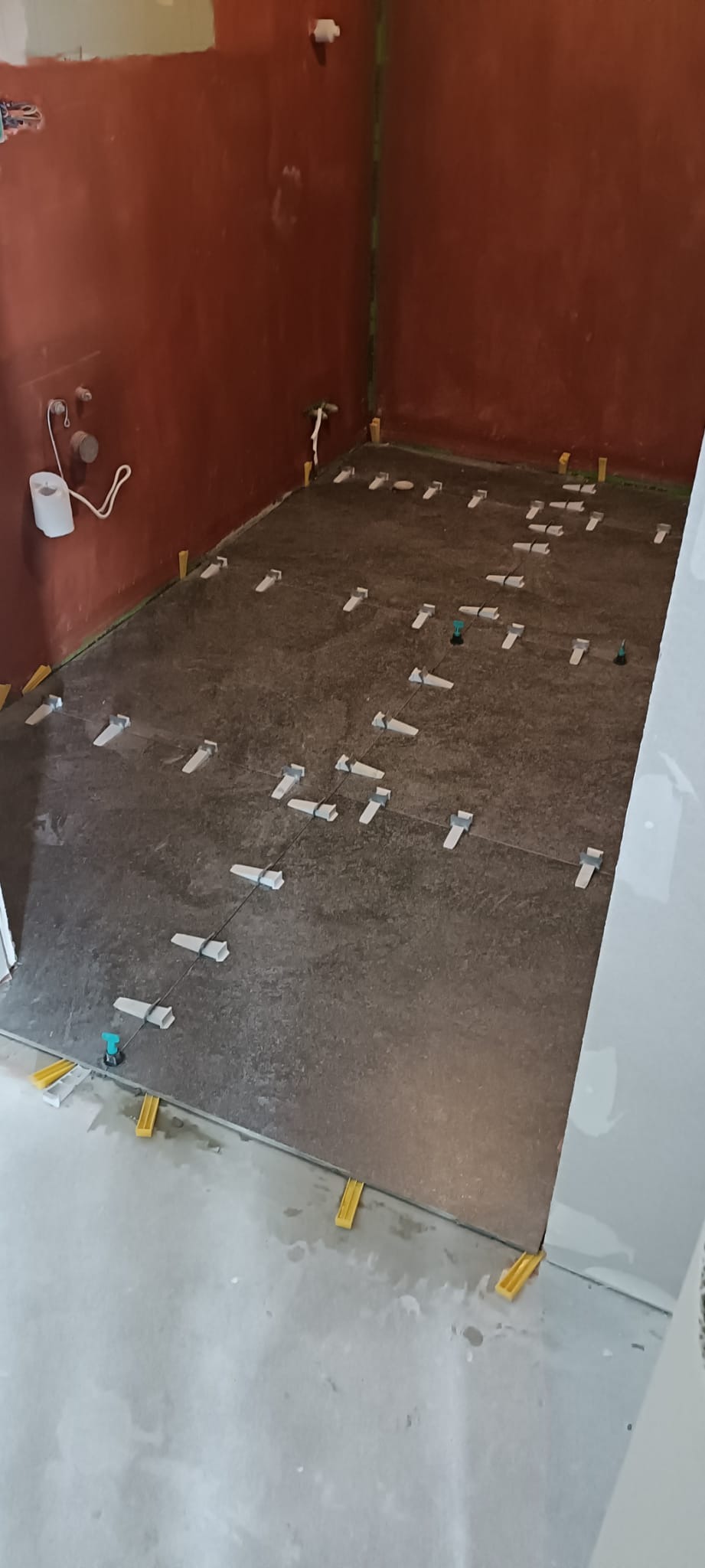 Układanie płytek podłogowych w łazience z użyciem systemu poziomowania. Widoczne płytki w odcieniach szarości, kliny i szczypce do poziomowania. Ściany w kolorze ceglastym.