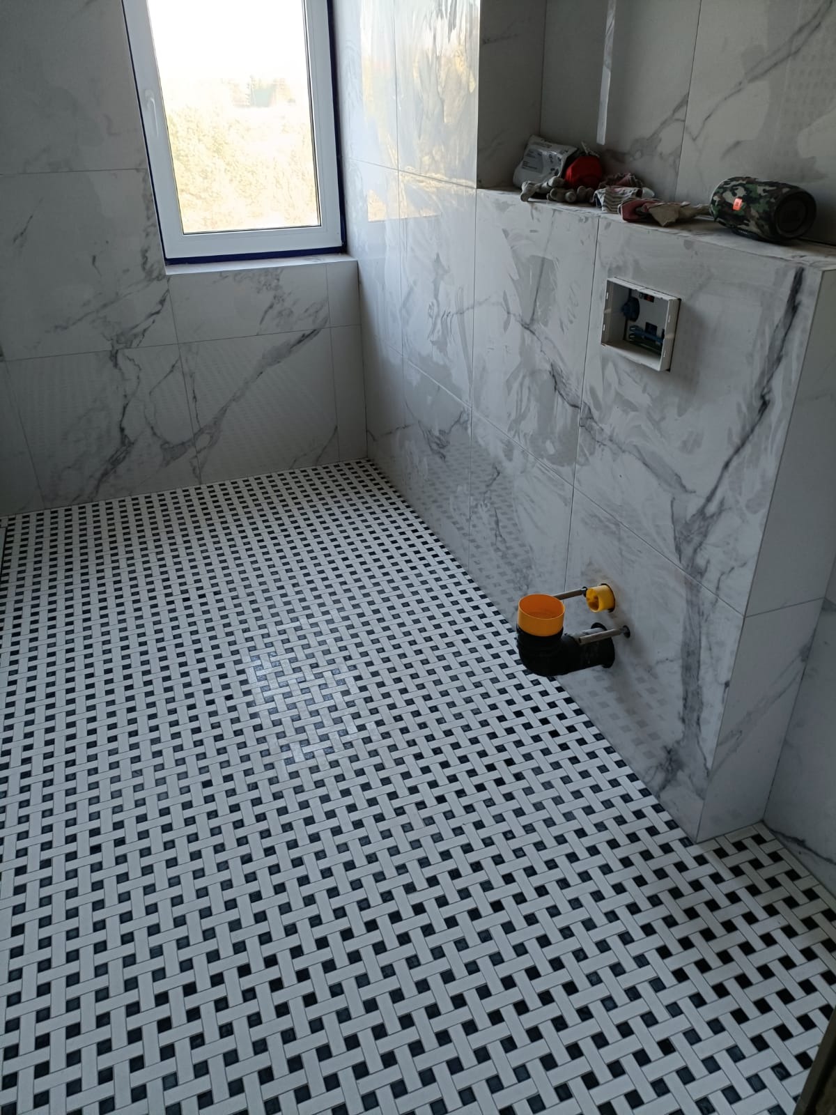 Wykończona łazienka z marmurowymi płytkami na ścianach i podłodze w czarno-białą kratkę. Widoczne elementy instalacji hydraulicznej i okno z niebieską ramą.