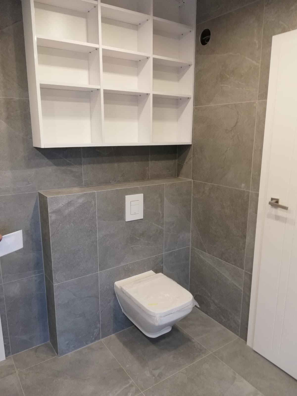 Nowoczesna łazienka w szarościach z podwieszaną toaletą, białą szafką i płytkami imitującymi kamień. Toaleta zabezpieczona folią ochronną.