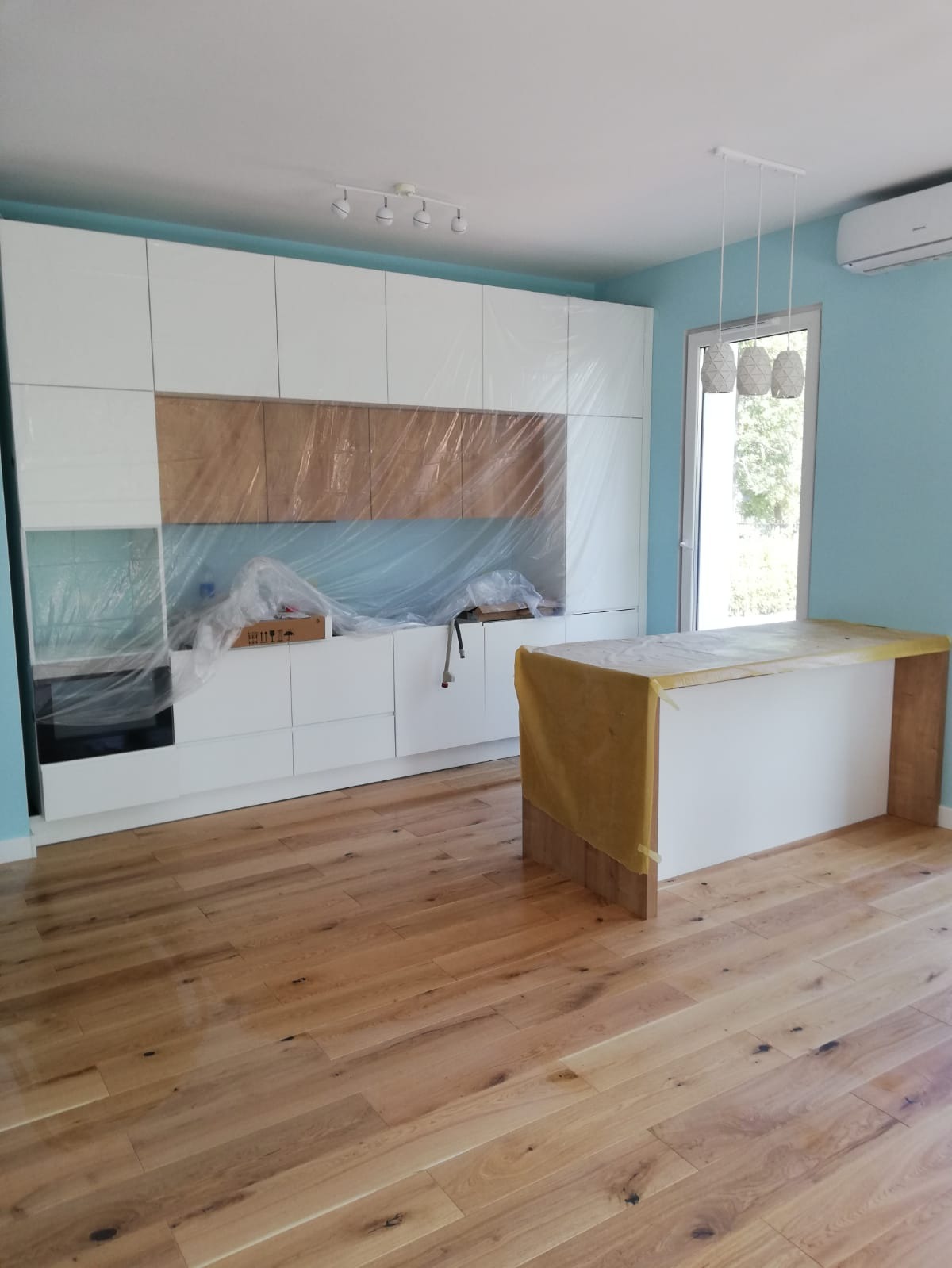 Jasna kuchnia w trakcie wykończenia: drewniana podłoga, białe szafki zabezpieczone folią, wyspa kuchenna z blatem osłoniętym kartonem. Ściany w odcieniach błękitu.