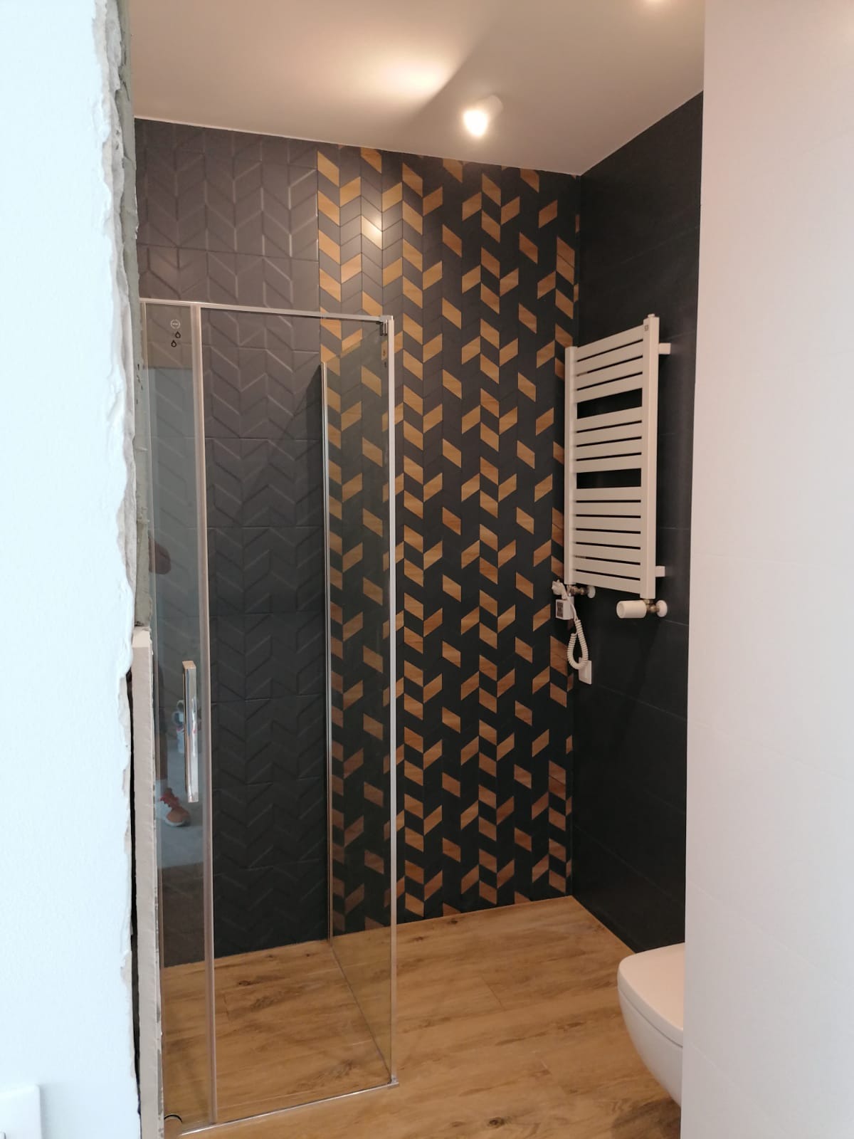Nowoczesna łazienka po remoncie: kabina prysznicowa ze szklanymi drzwiami, dekoracyjne płytki w jodełkę, grzejnik drabinkowy i drewnopodobna podłoga.