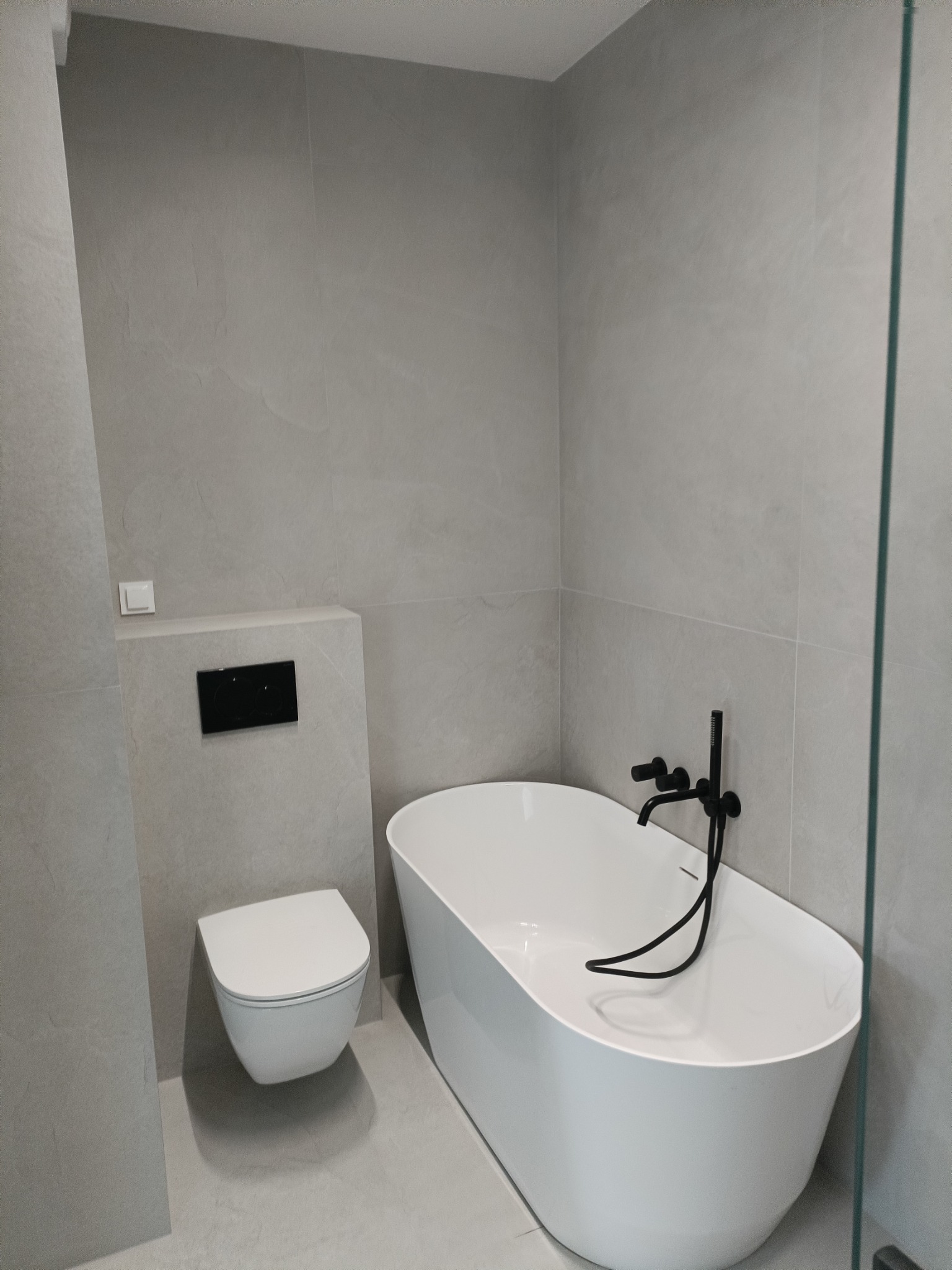 Minimalistyczna łazienka w szarościach z wolnostojącą wanną i podwieszaną toaletą. Czarna armatura i szklana kabina prysznicowa dodają elegancji.