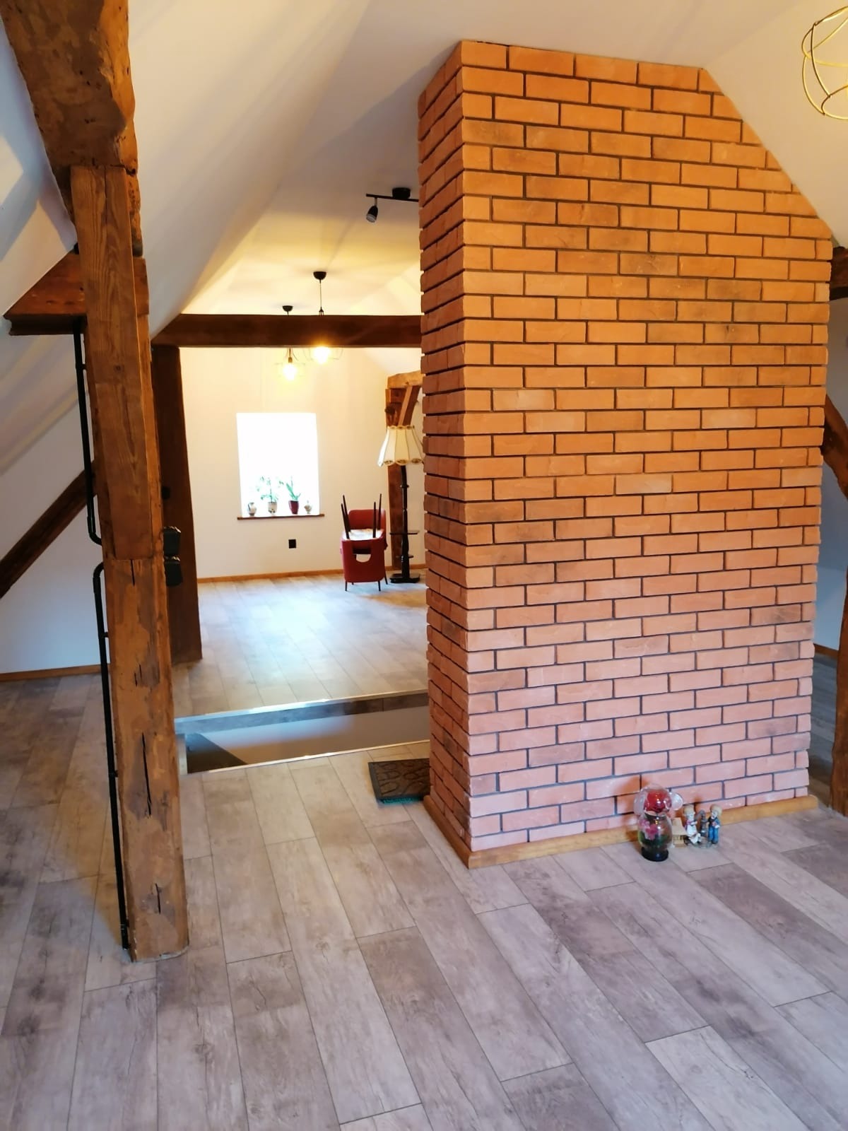 Poddasze po remoncie: ceglana kolumna, drewniane belki, podłoga imitująca drewno. Widok na jasne pomieszczenie z oknem i czerwonym krzesłem. Dekoracje na podłodze.