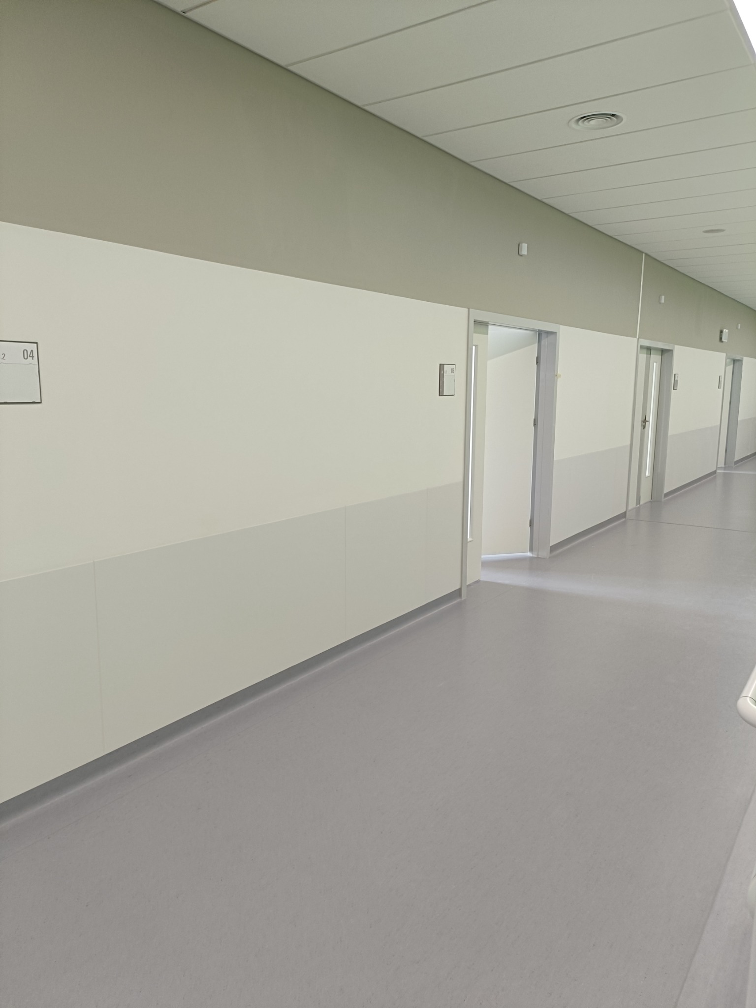 Długi, jasny korytarz biurowy z szarymi panelami ściennymi i podłogą. Widoczne drzwi i numeracja pomieszczeń. Minimalistyczny design, neutralna kolorystyka.