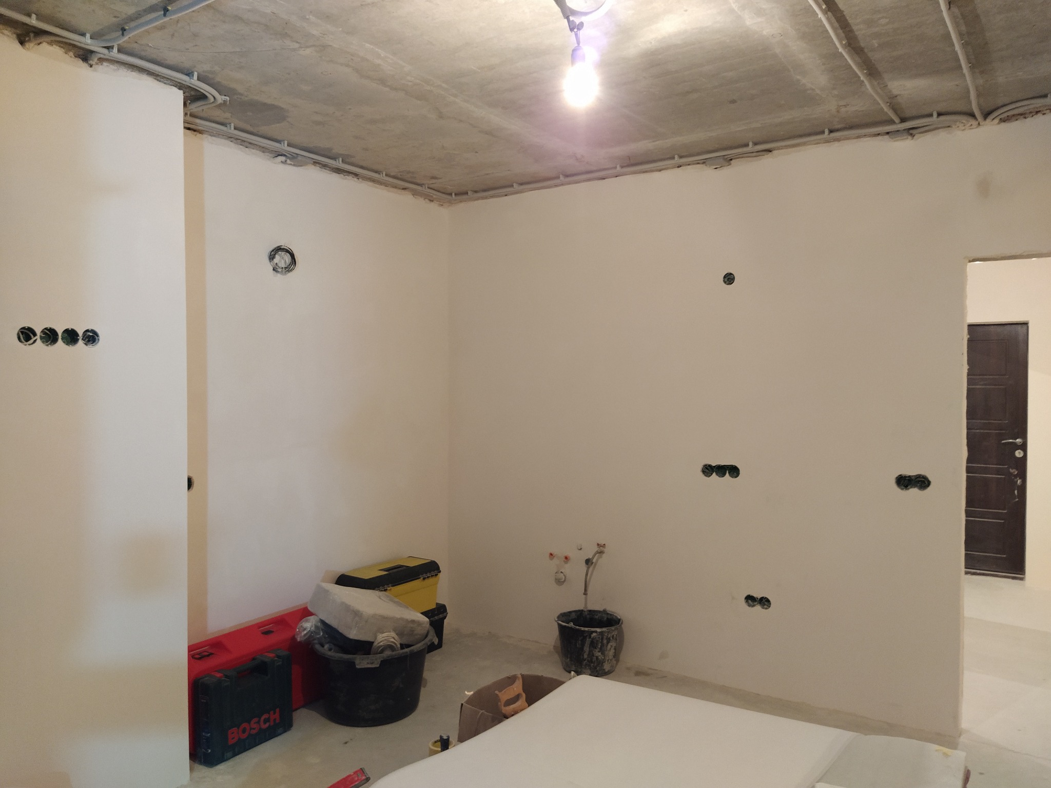 Wnętrze w trakcie remontu: surowy betonowy sufit z instalacją elektryczną, świeżo pomalowane ściany z puszkami elektrycznymi, narzędzia Bosch w tle.