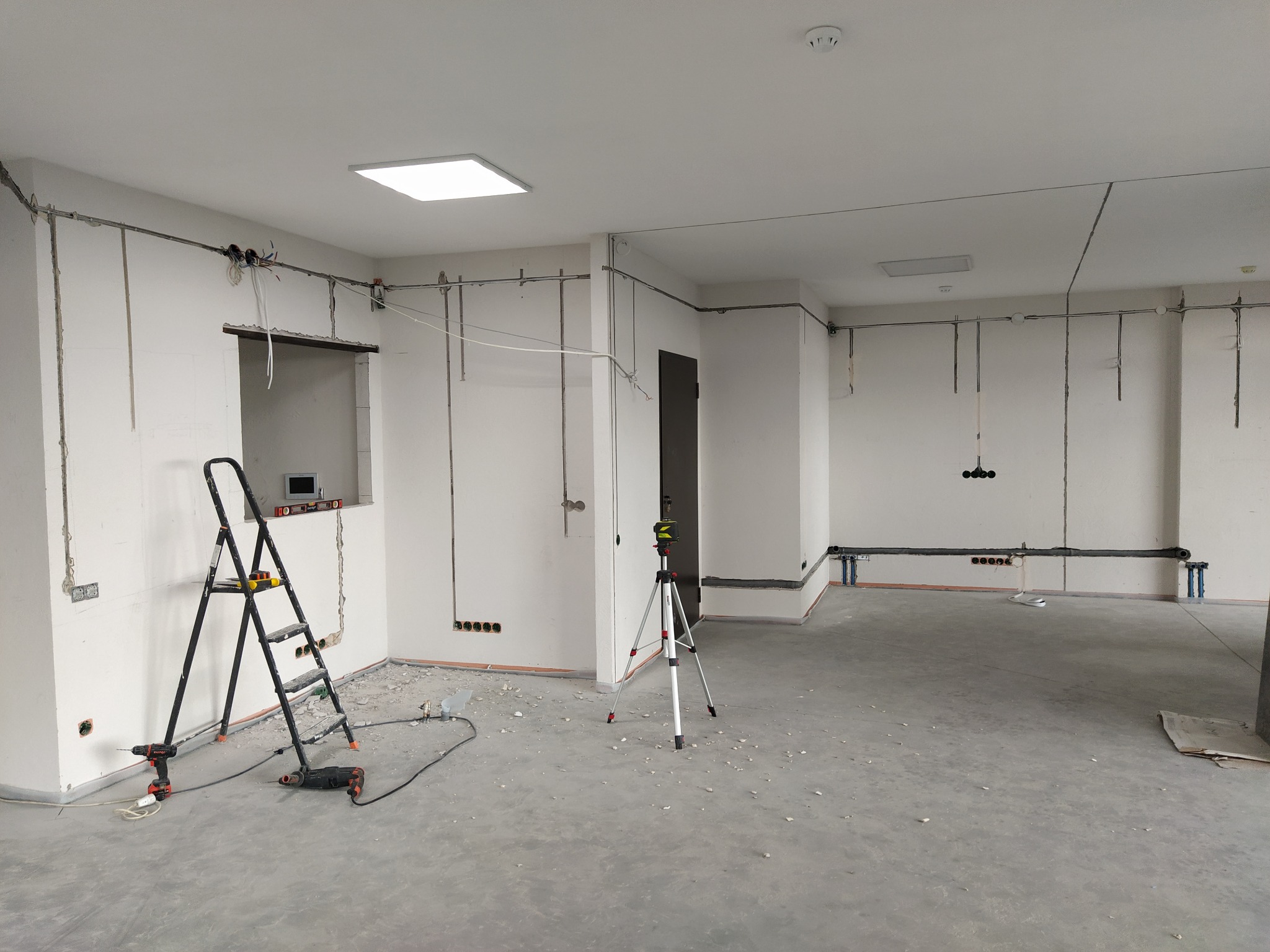 Wnętrze w trakcie prac elektrycznych: przewody, puszki, drabina i poziomica laserowa. Widoczne ślady po kuciu w ścianach. Surowy beton na podłodze.