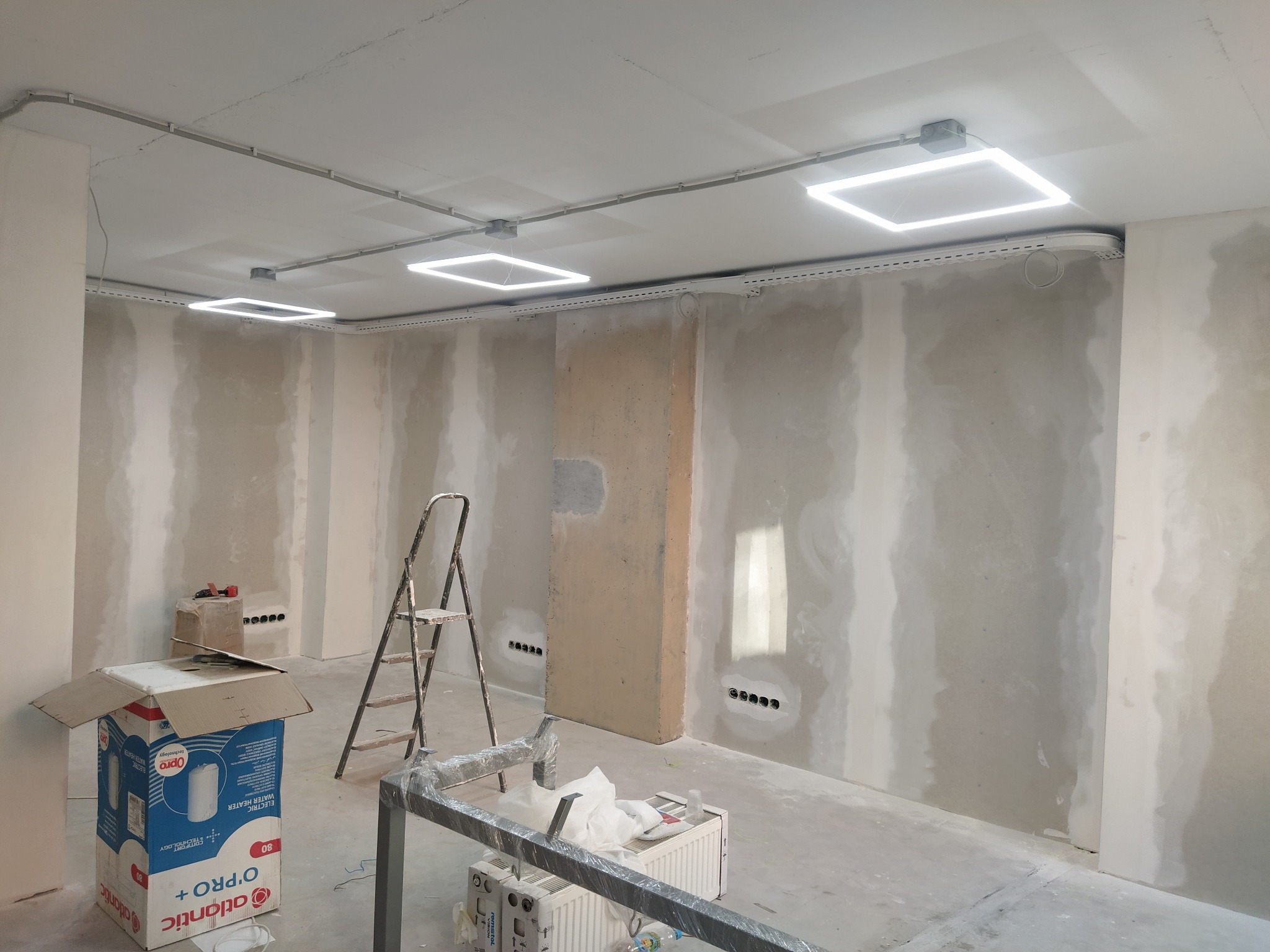 Wnętrze w trakcie prac wykończeniowych: ściany z gładzią, sufit z kwadratowymi lampami LED, widoczne kable i puszki elektryczne, drabina, karton po bojlerze.